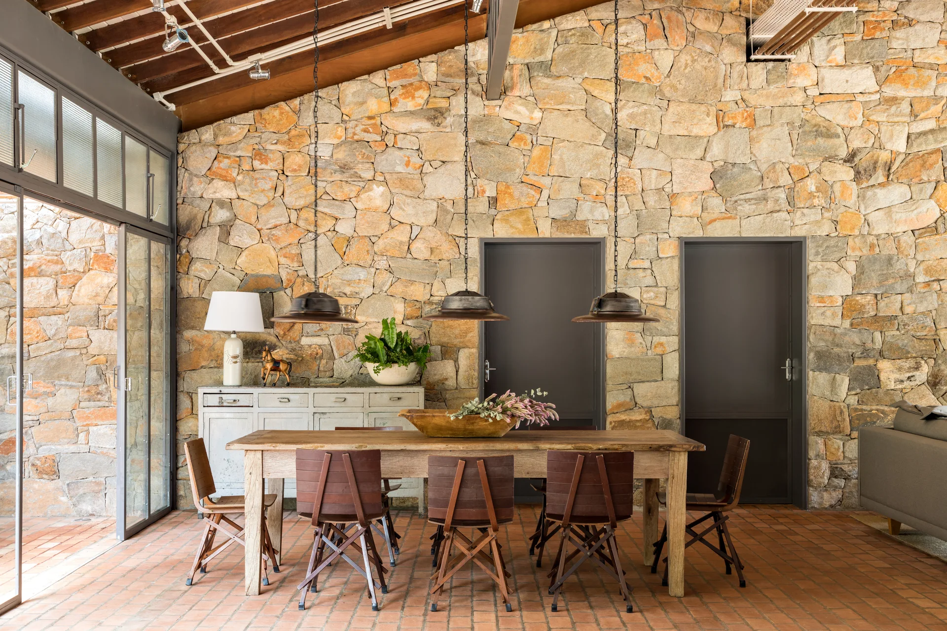 A rustic dining area features a wooden table and chairs set against a stone wall with three industrial pendant lights hanging from a vaulted ceiling.