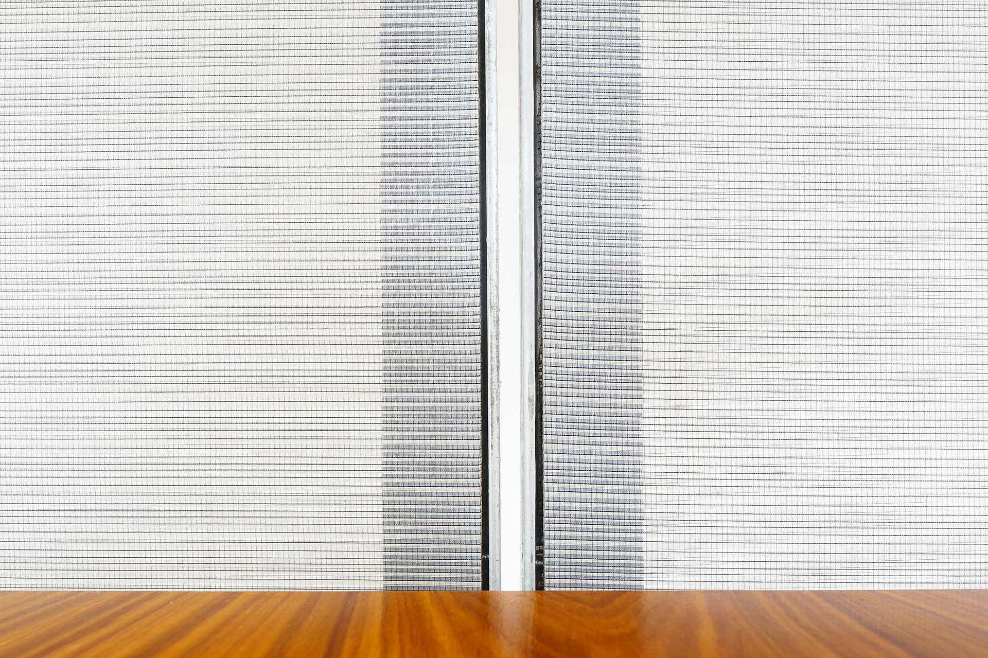A polished wood tabletop sits in front of textured grey mesh panels divided by a thin vertical frame.