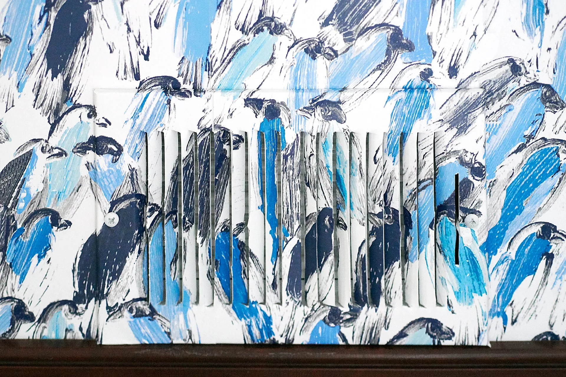 A close-up of a blue and white bird patterned wallpaper with a metal louvered air vent painted to match the illustration.