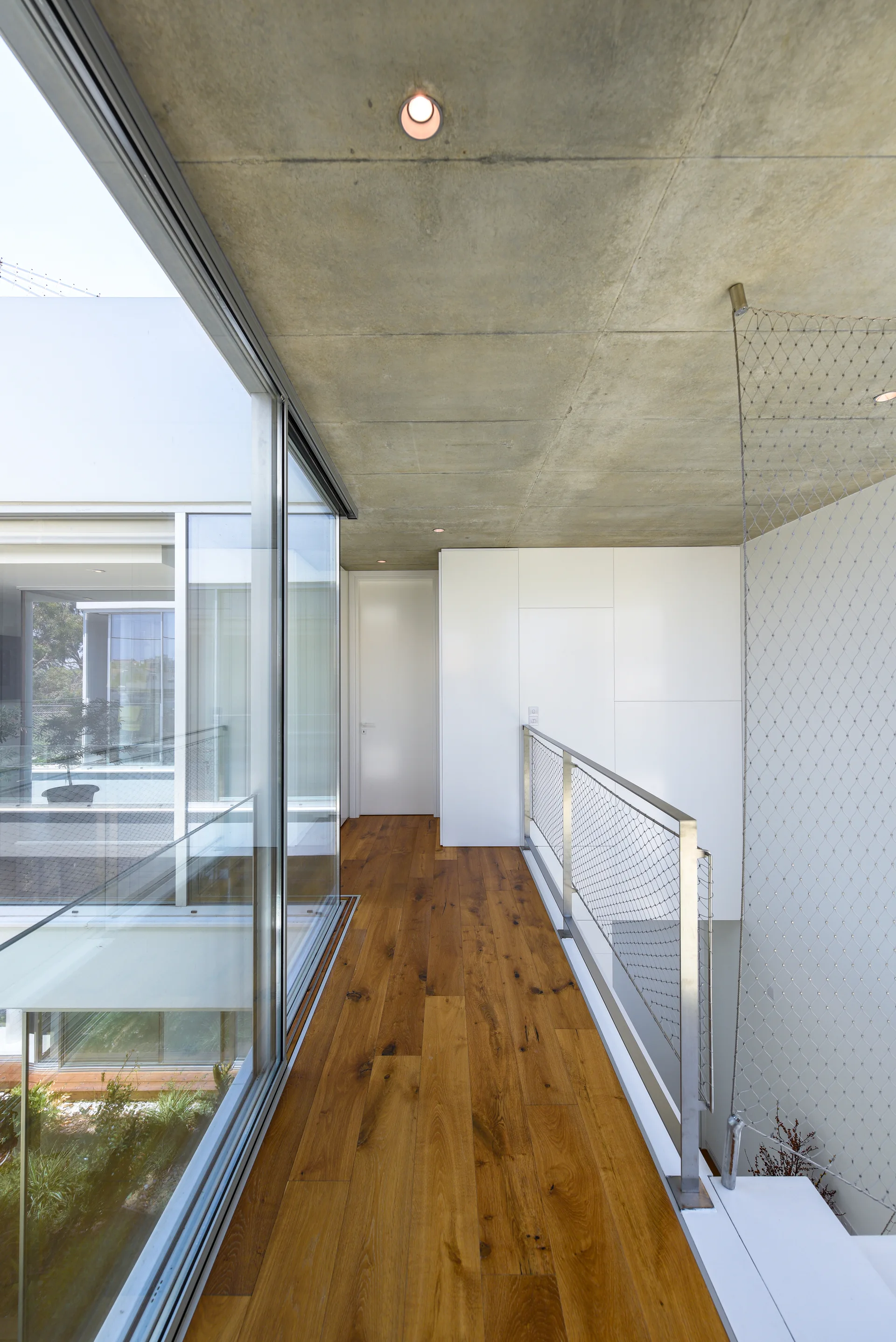 A minimalist hallway features medium-brown wood flooring, an exposed concrete ceiling, a metal mesh railing, and a floor-to-ceiling glass wall.