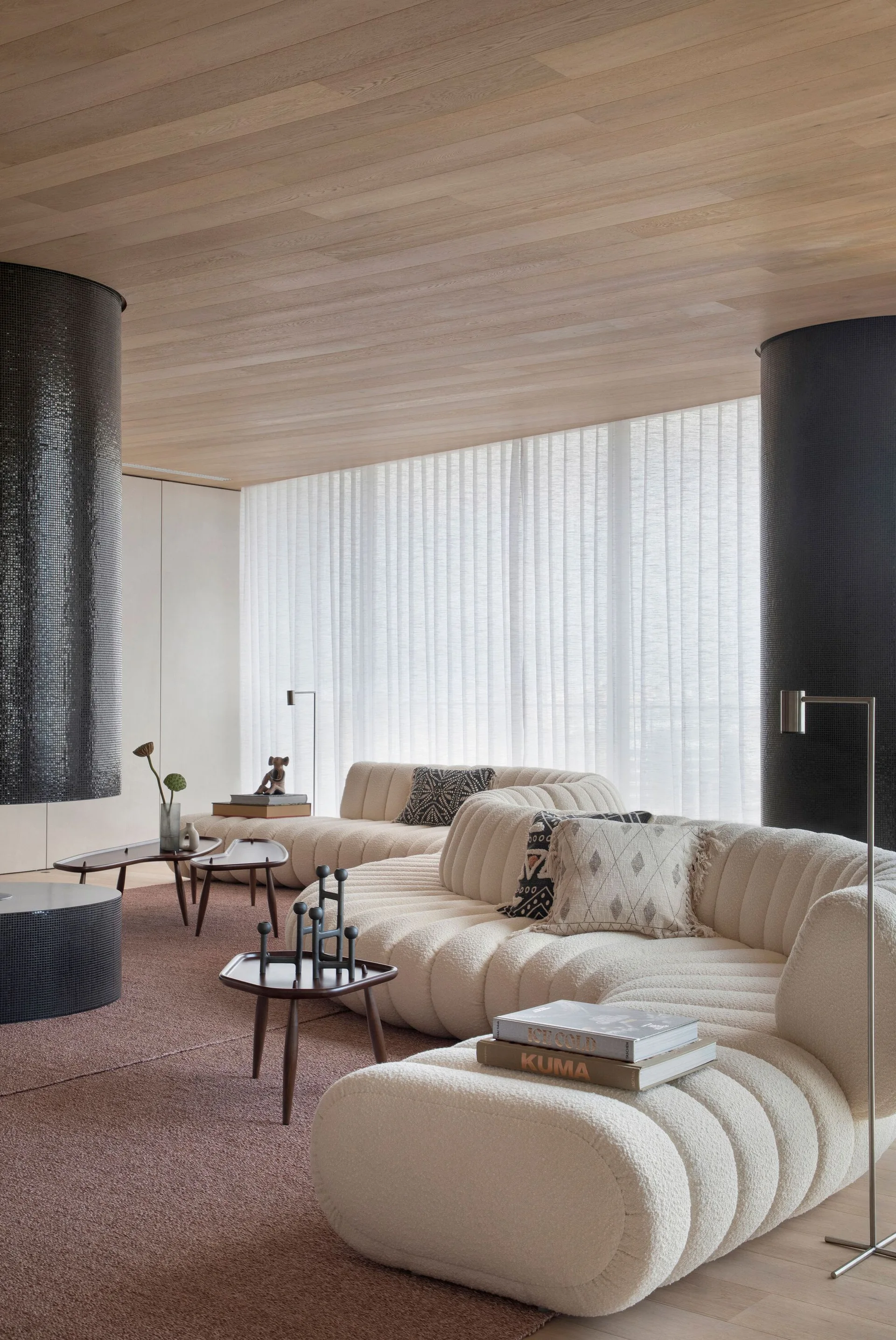 A contemporary living room featuring a curved cream-colored sofa, low wooden tables, and black tiled columns beneath a light wood plank ceiling.