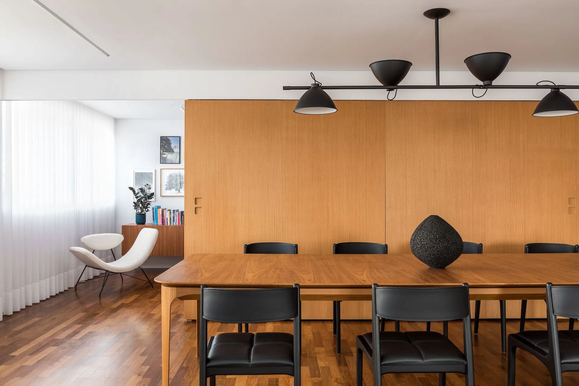 A wooden dining table with black leather-like chairs sits in front of a large light wood sliding partition wall beneath a linear black light fixture.