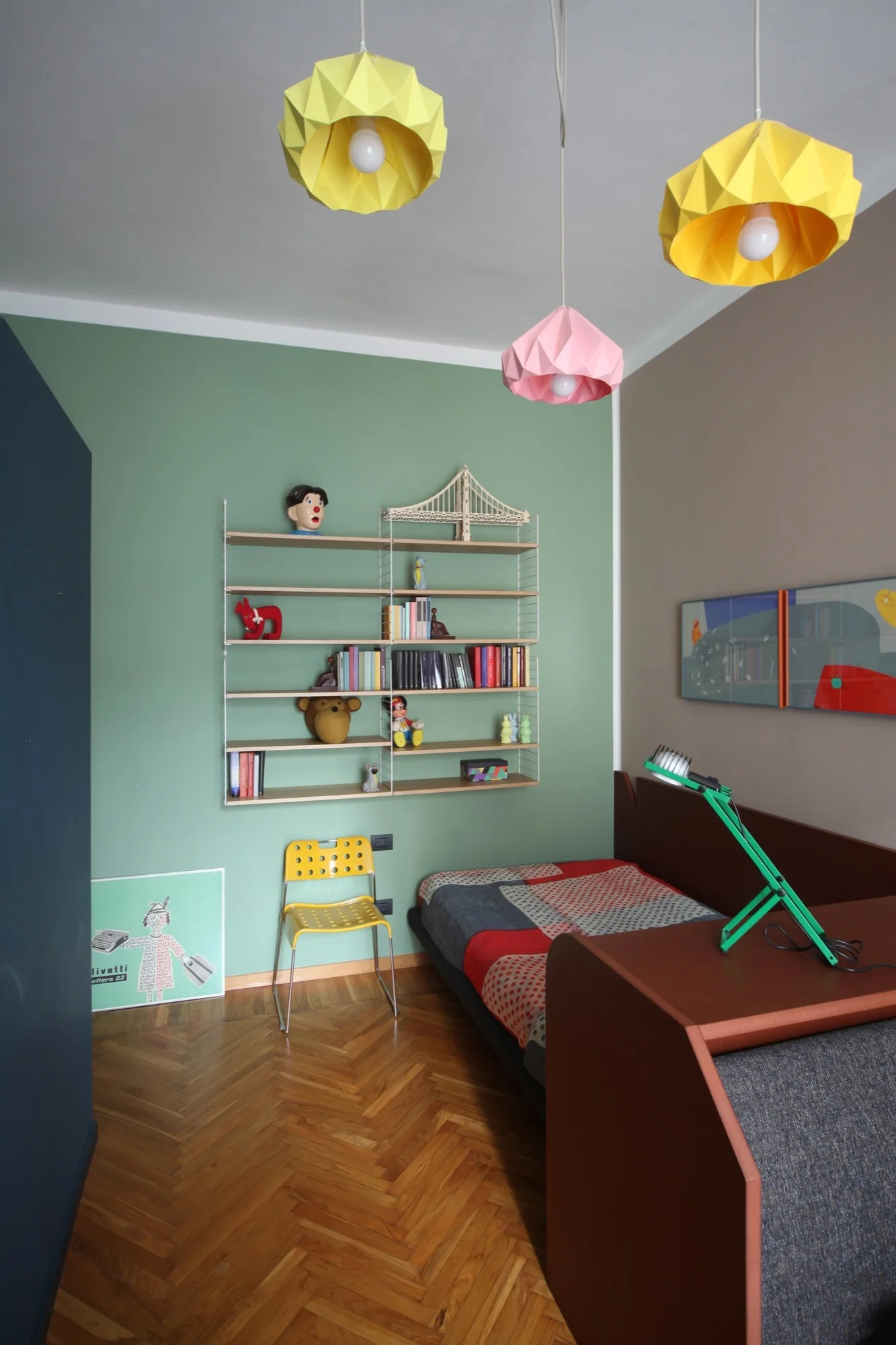 A child's bedroom features a sage green wall with modular shelving, a low bed, a yellow chair, and origami-style pendant lights.