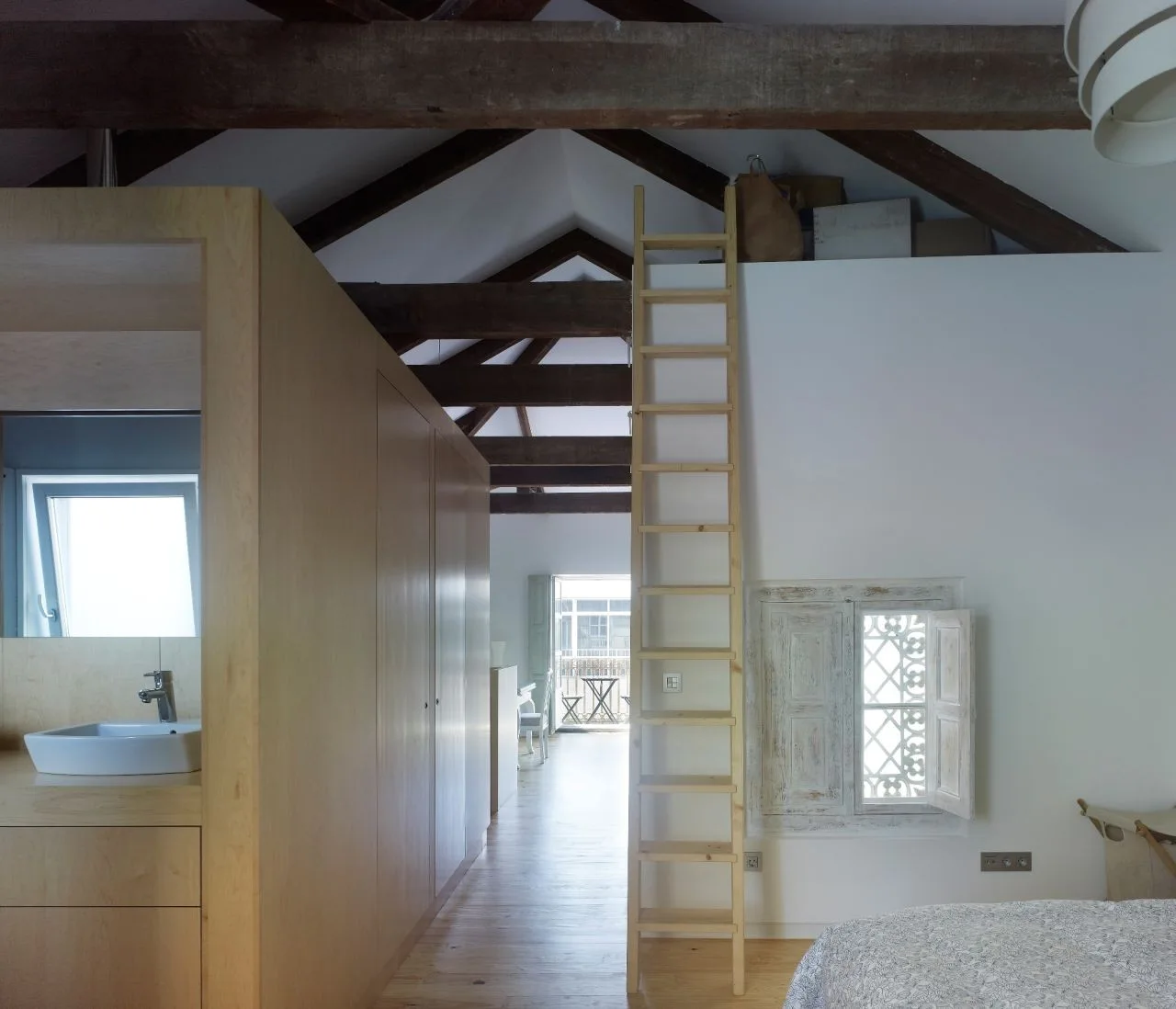 A loft bedroom featuring dark exposed ceiling beams, a large light-wood wardrobe partition, an integrated vessel sink, and a tall wooden ladder.