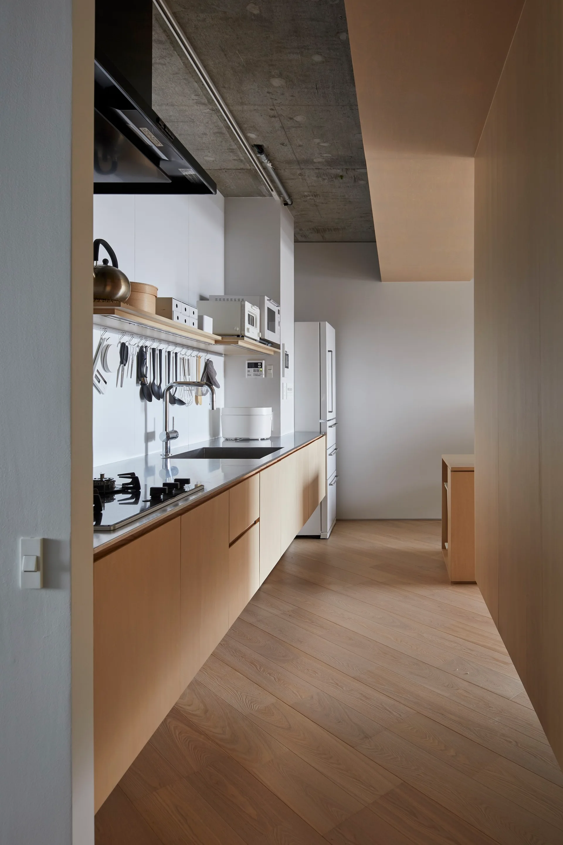 Minimalist galley kitchen with light wood cabinetry, a long stainless steel countertop, and a raw concrete ceiling above diagonal wood flooring.