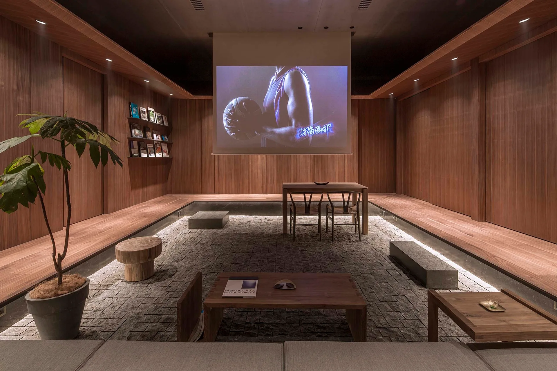 A minimalist media room features a sunken cobblestone floor surrounded by warm wood-paneled walls, low wooden tables, and a large projector screen.