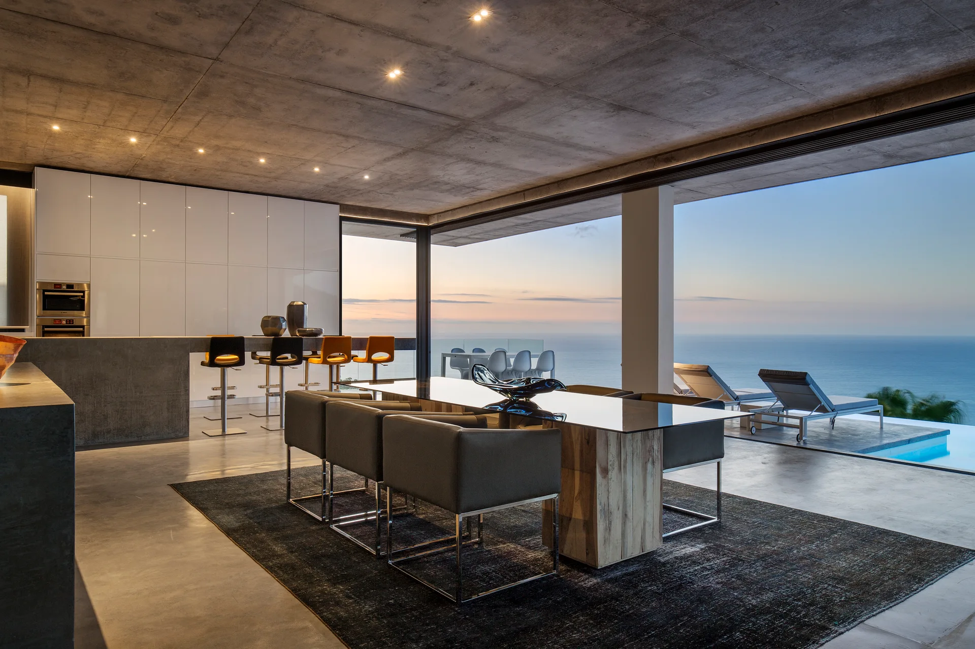 A modern open-concept dining area features a glass-top table on a dark rug beneath an exposed concrete ceiling looking out to an oceanfront pool.