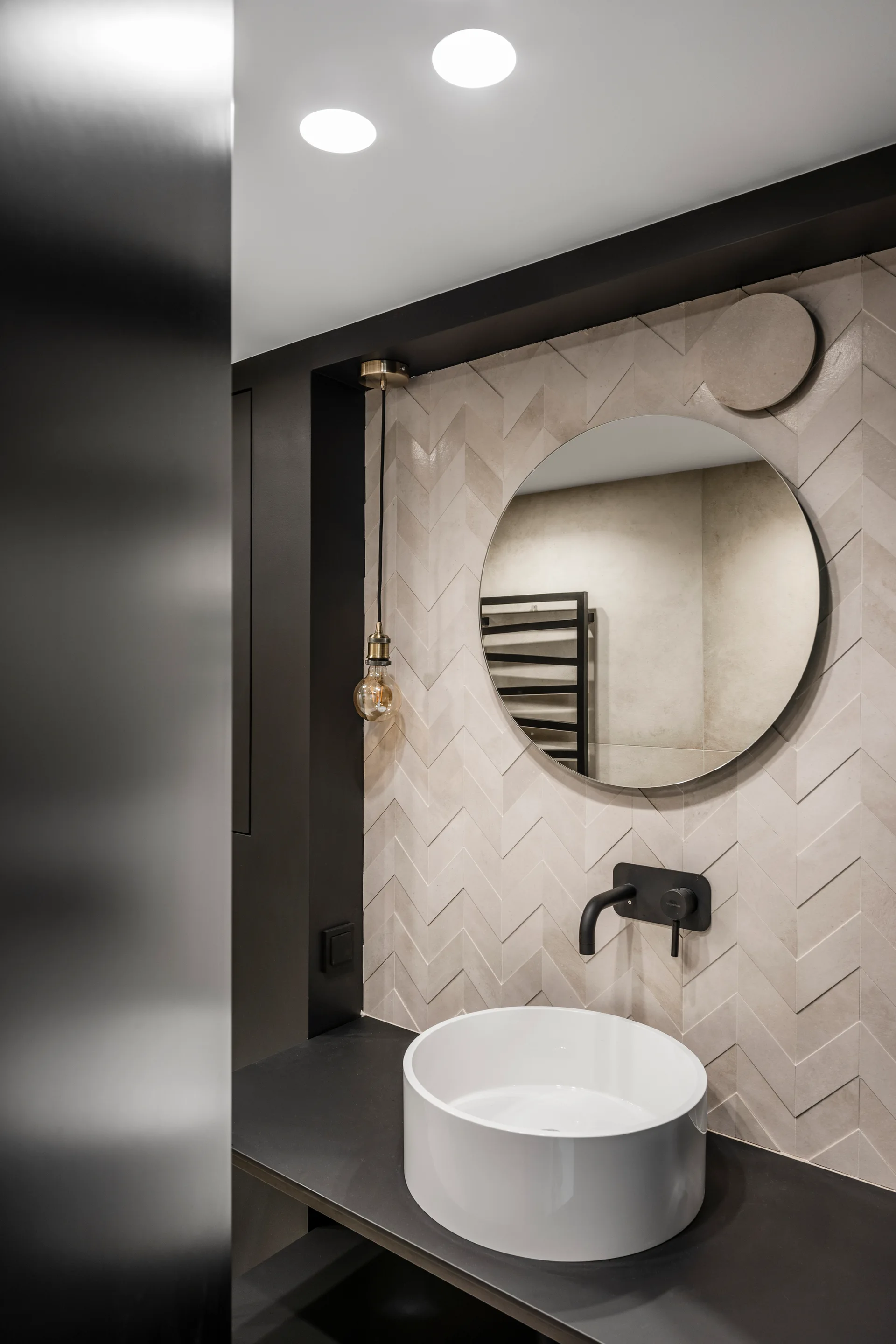 A modern bathroom vanity features a round white vessel sink on a black countertop, set against a beige herringbone tile backsplash with a circular mirror.