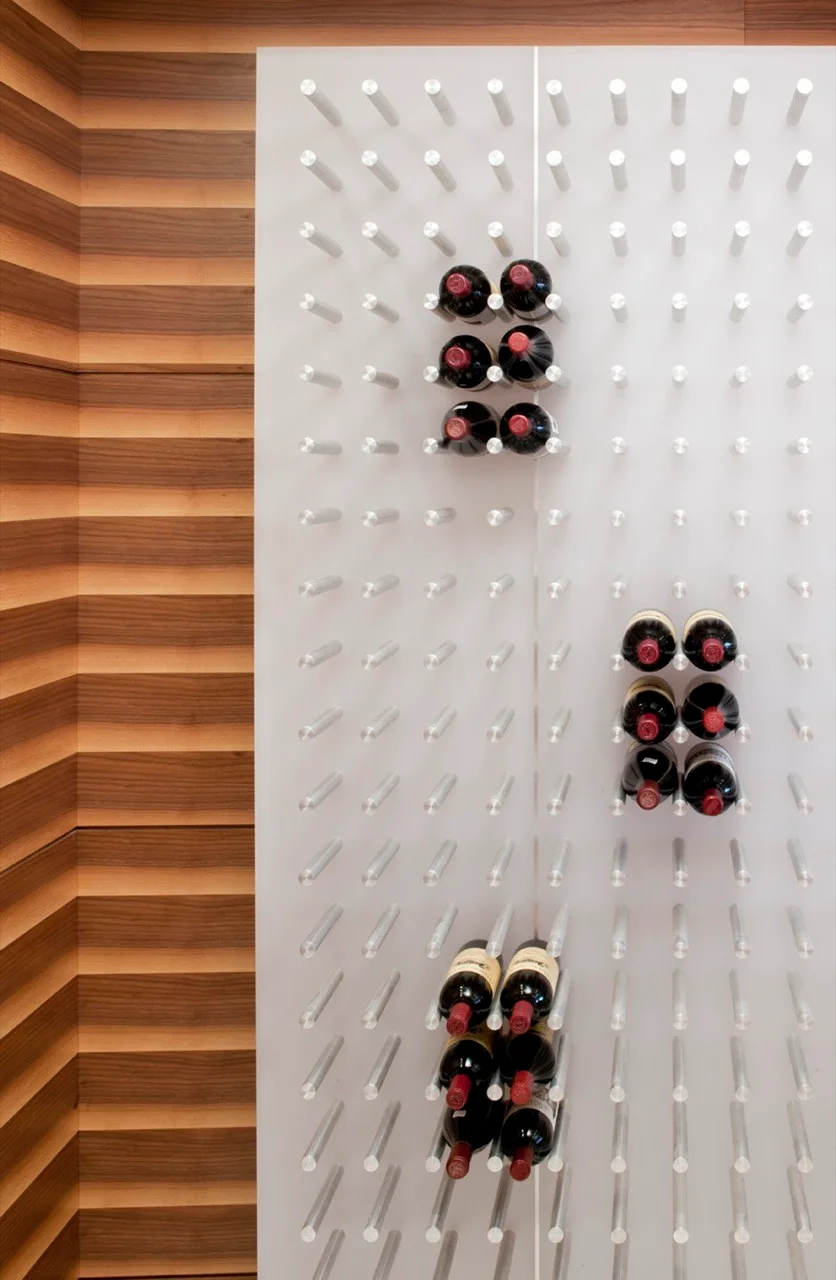 A minimalist wall-mounted wine rack with white backing and metal pegs displays bottles horizontally against a slatted wood panel background.