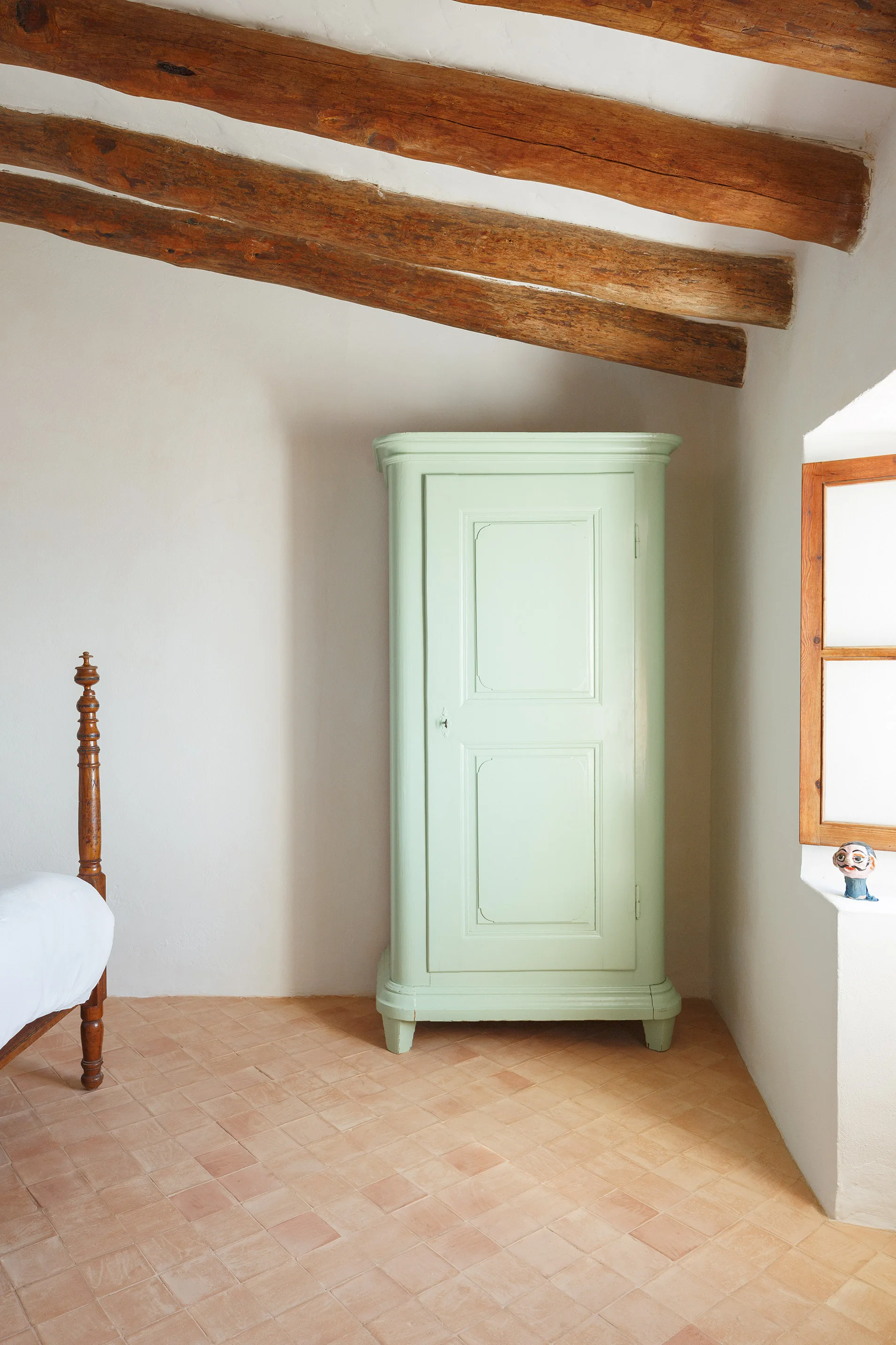A rustic bedroom corner features a pale green wooden wardrobe, terracotta tile floors, and exposed wooden beams across the ceiling.