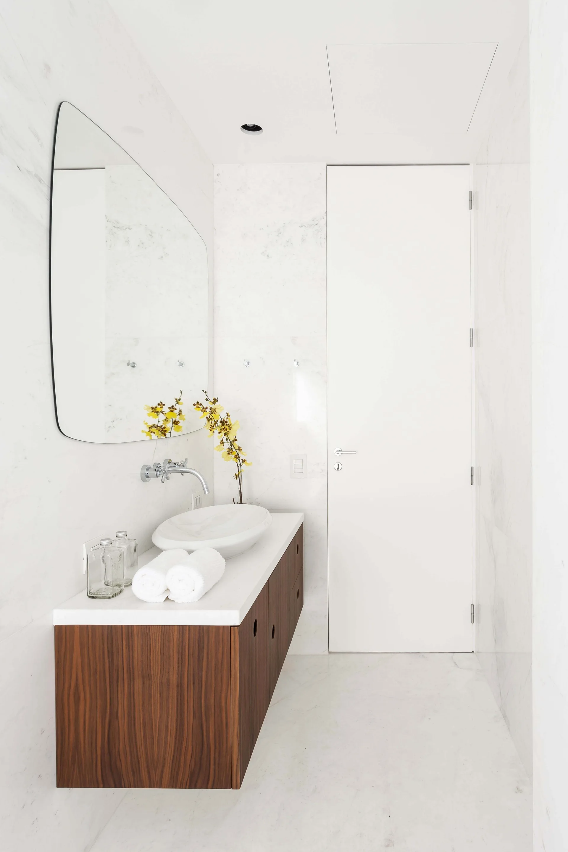 A minimalist bathroom features a floating wood vanity with a white vessel sink, a chrome faucet, and a large rounded mirror against marble walls.