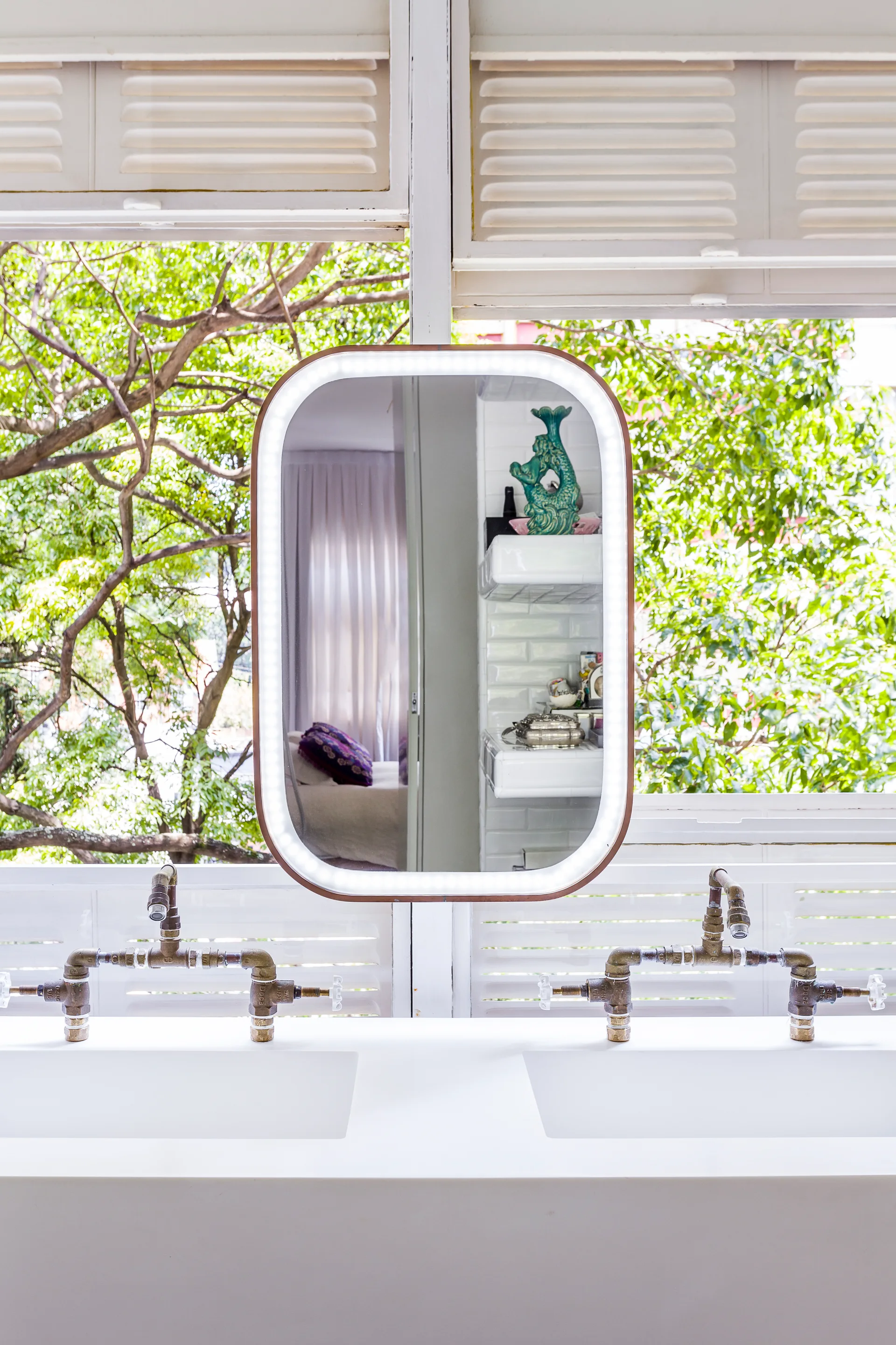 A white double vanity features two brass industrial pipe faucets and a central rounded lighted mirror set against white shuttered windows.
