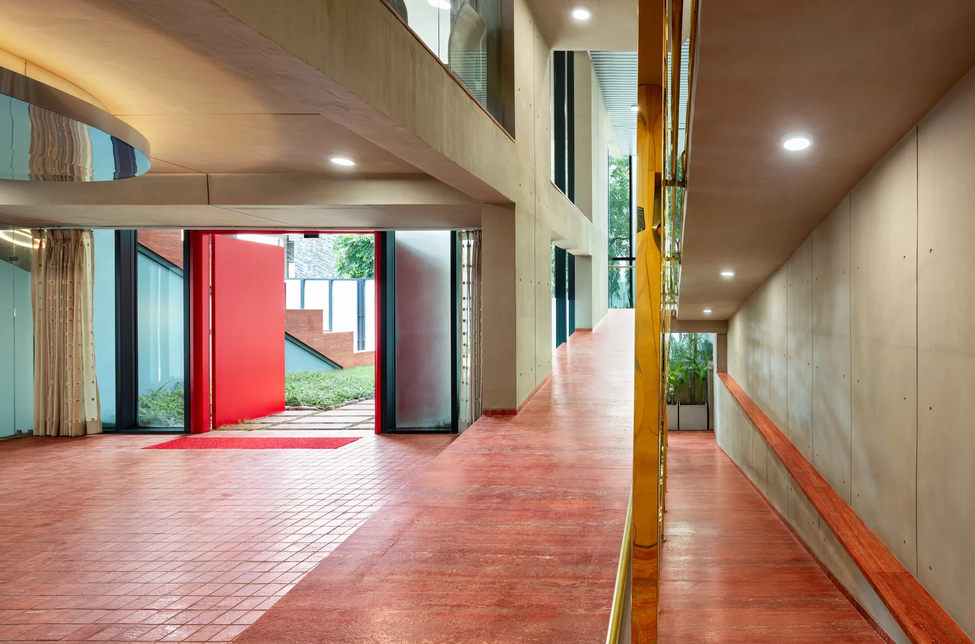 A modern brutalist entryway featuring raw concrete walls, a large open red door, and a sloping red ramp with a gold railing.