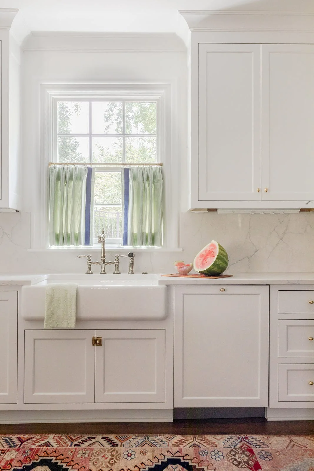 A white farmhouse kitchen sink area featuring a polished bridge faucet, striped green cafe curtains, shaker cabinetry, and a patterned rug.