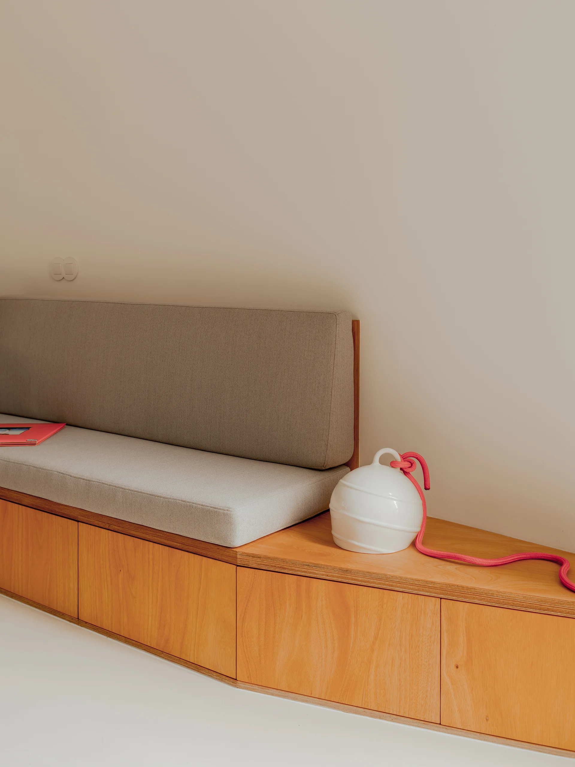 A minimalist built-in wooden bench with a light grey cushion features integrated storage cabinets below and a white ceramic vessel on its right side.
