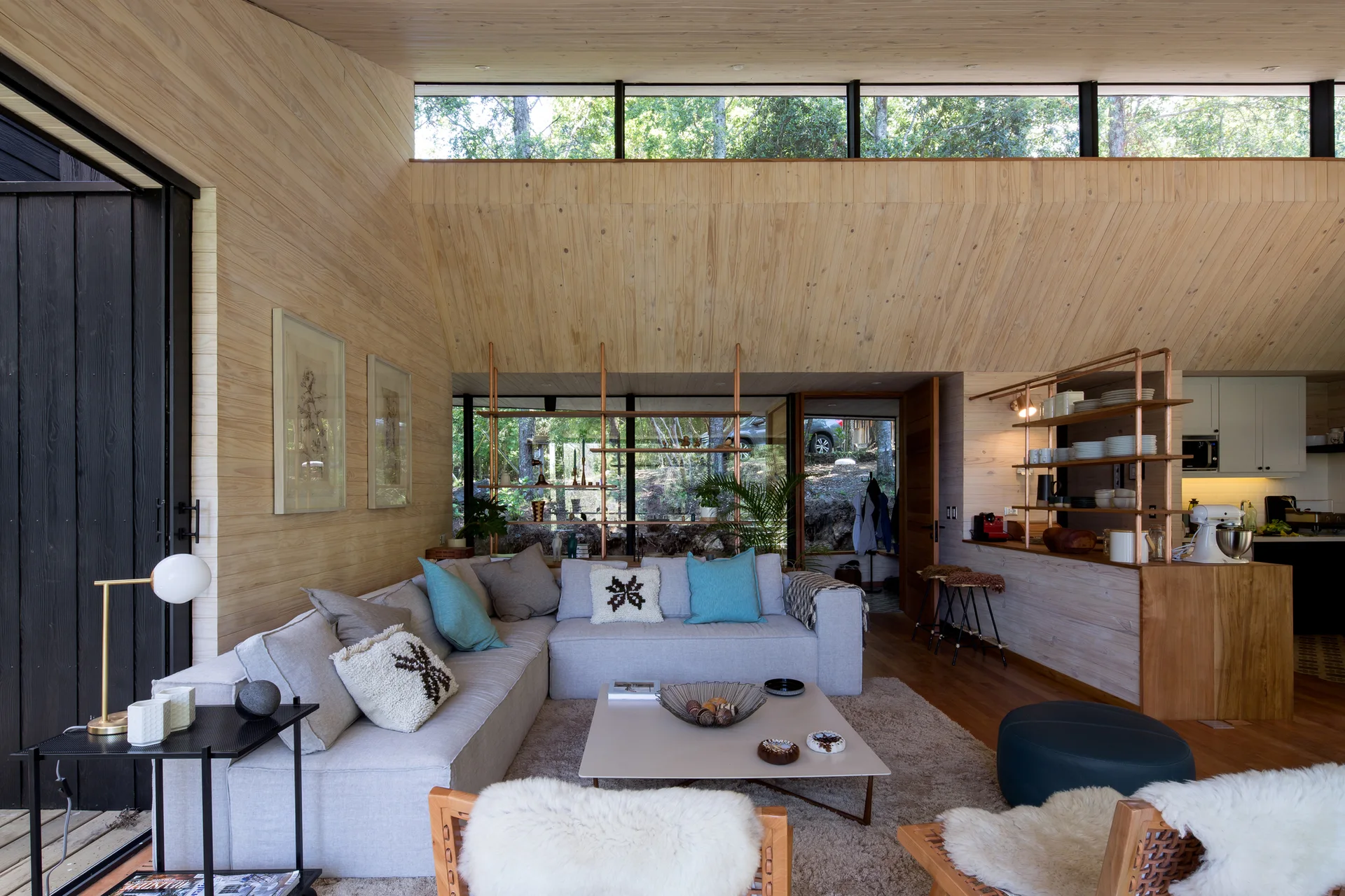 A modern living room features light wood walls and ceilings, a grey sectional sofa, and a custom copper pipe shelving unit dividing the space.