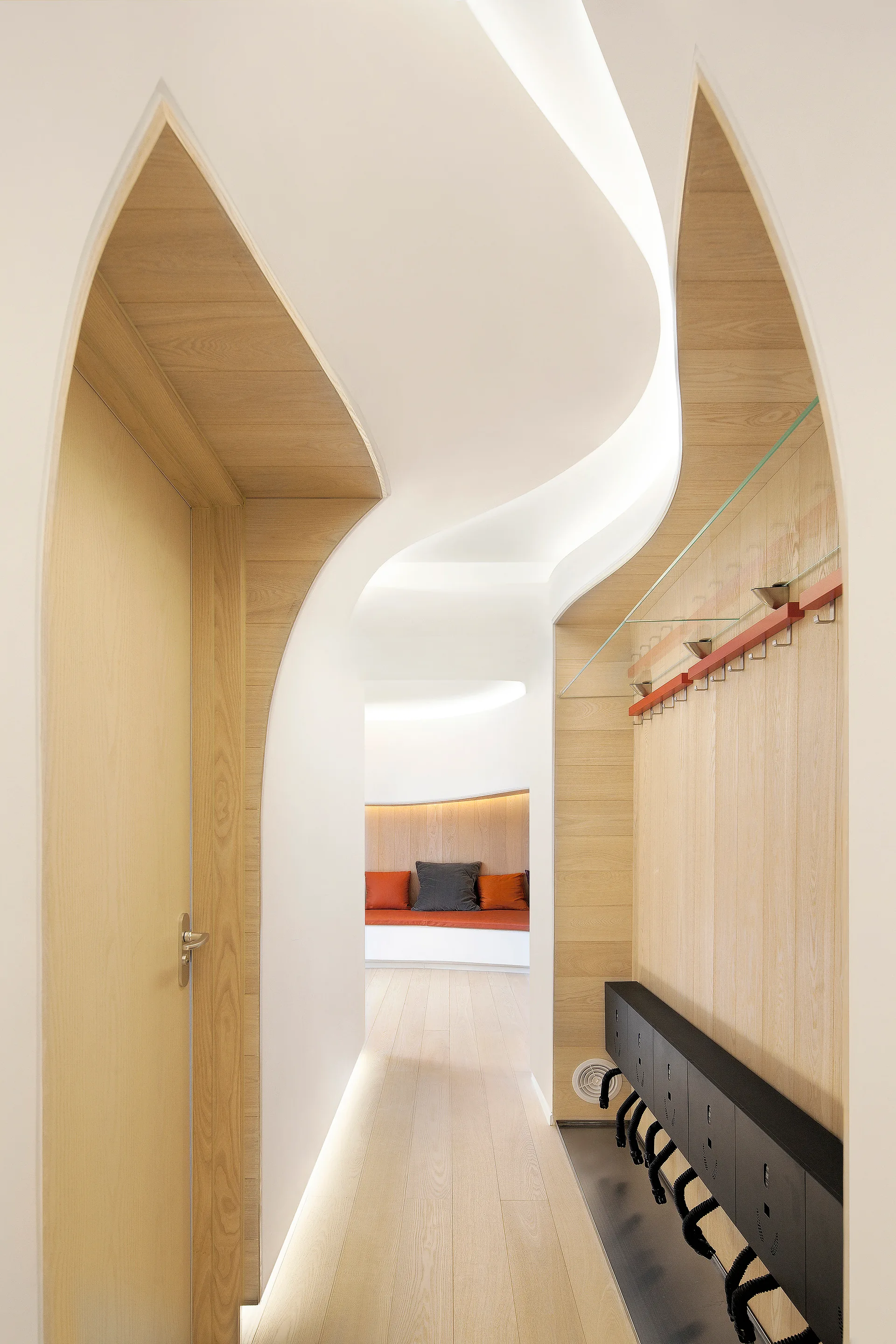A modern hallway featuring curved white plaster walls, integrated light wood paneling, recessed ceiling cove lighting, and a bench.