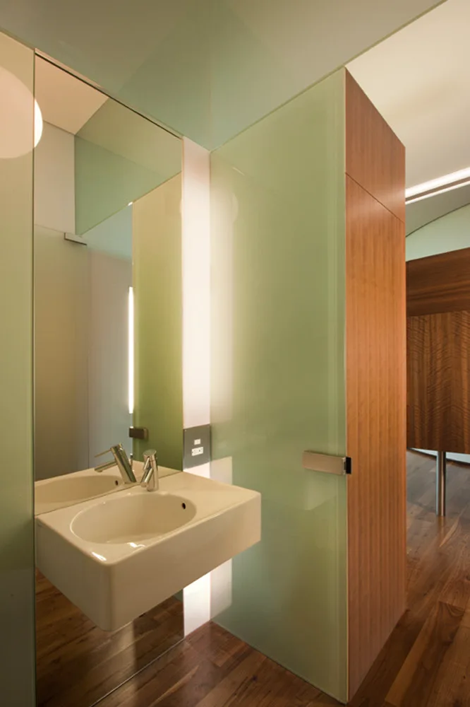 A modern bathroom features a wall-mounted white ceramic sink, a vertical mirror, green glass partition panels, and warm vertical wood cabinetry.