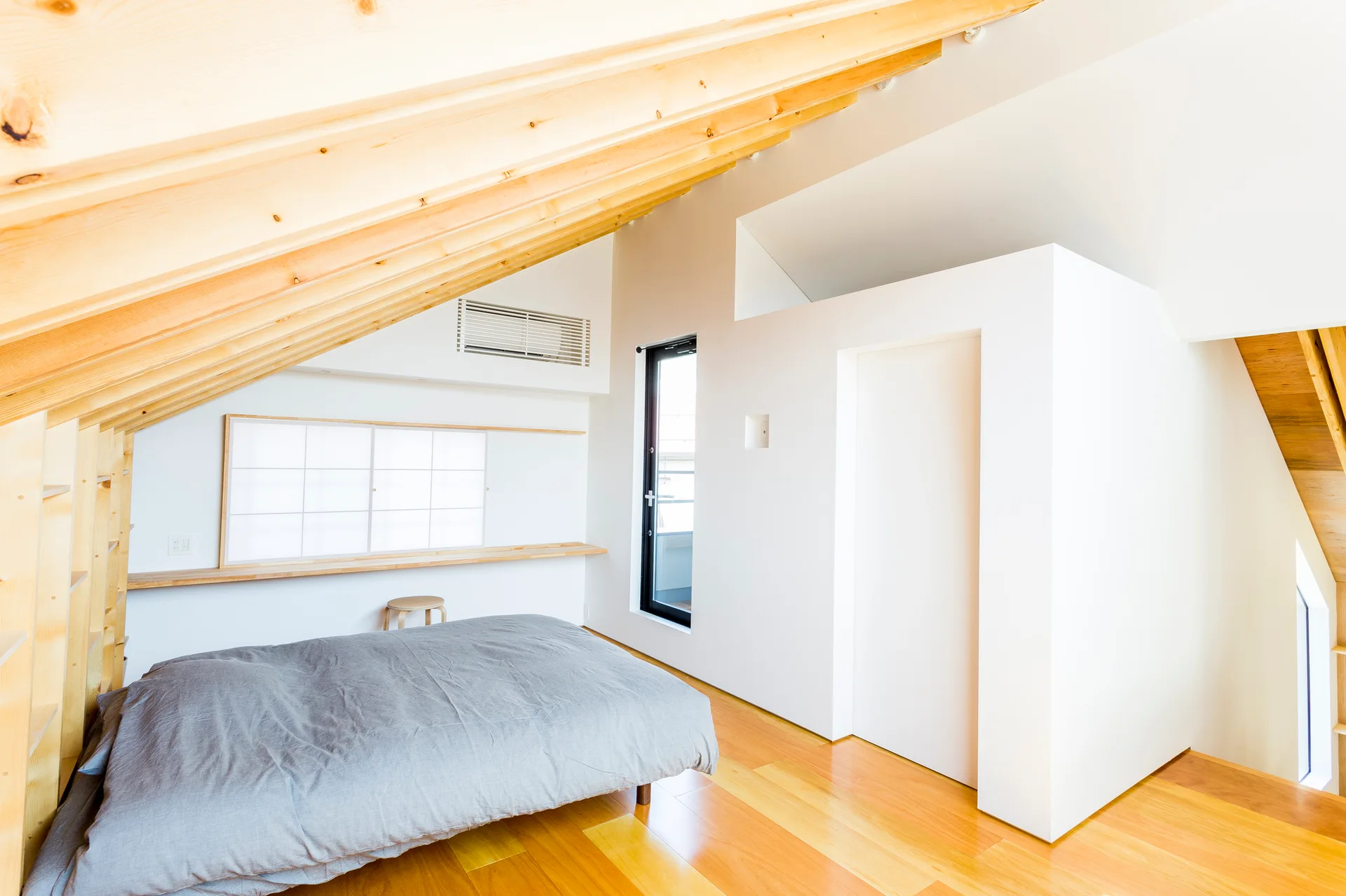 Minimalist attic bedroom with a low grey bed, sloped light wood ceiling beams, a long built-in wooden shelf, and white walls.