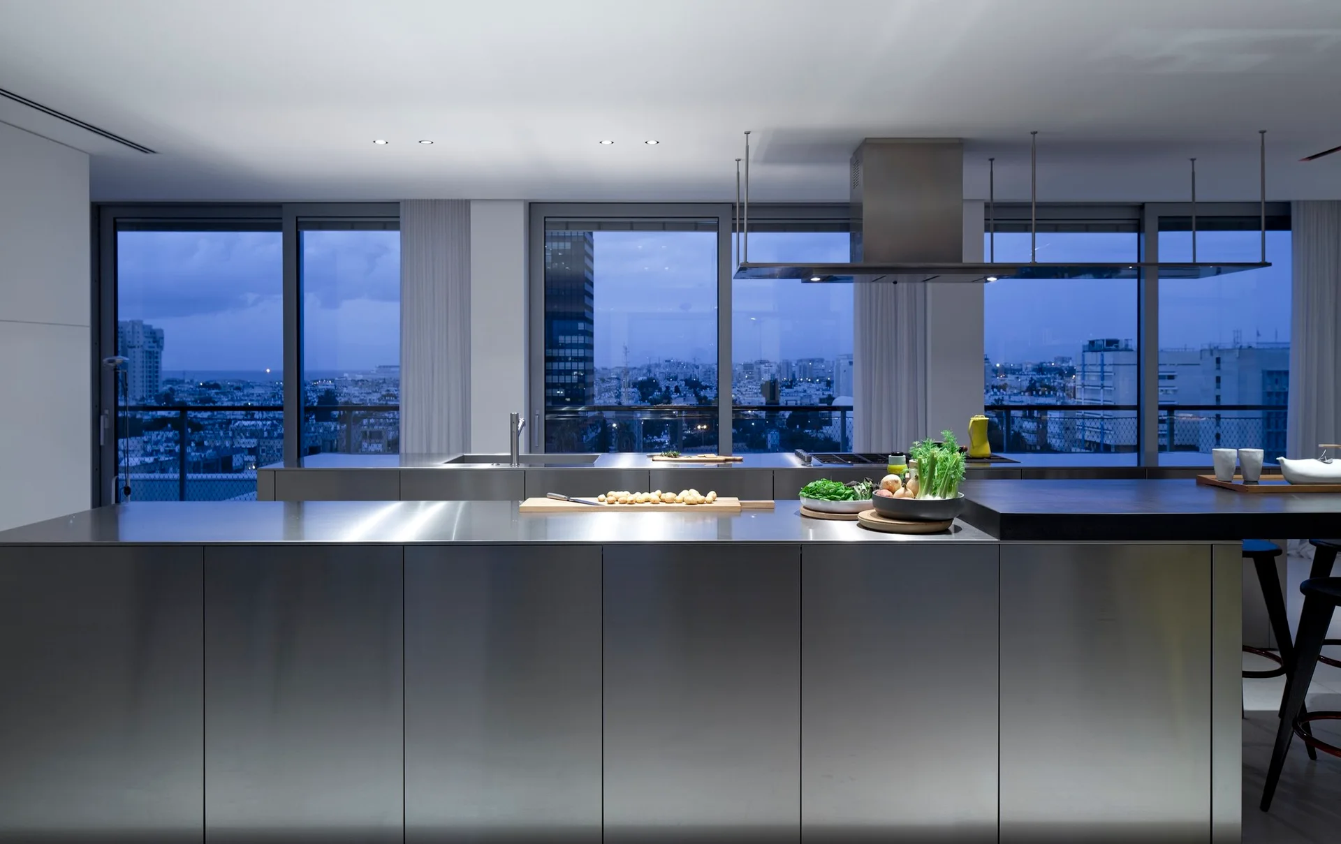 A modern kitchen featuring a brushed stainless steel island and matching range hood in front of large windows showing a city skyline at dusk.