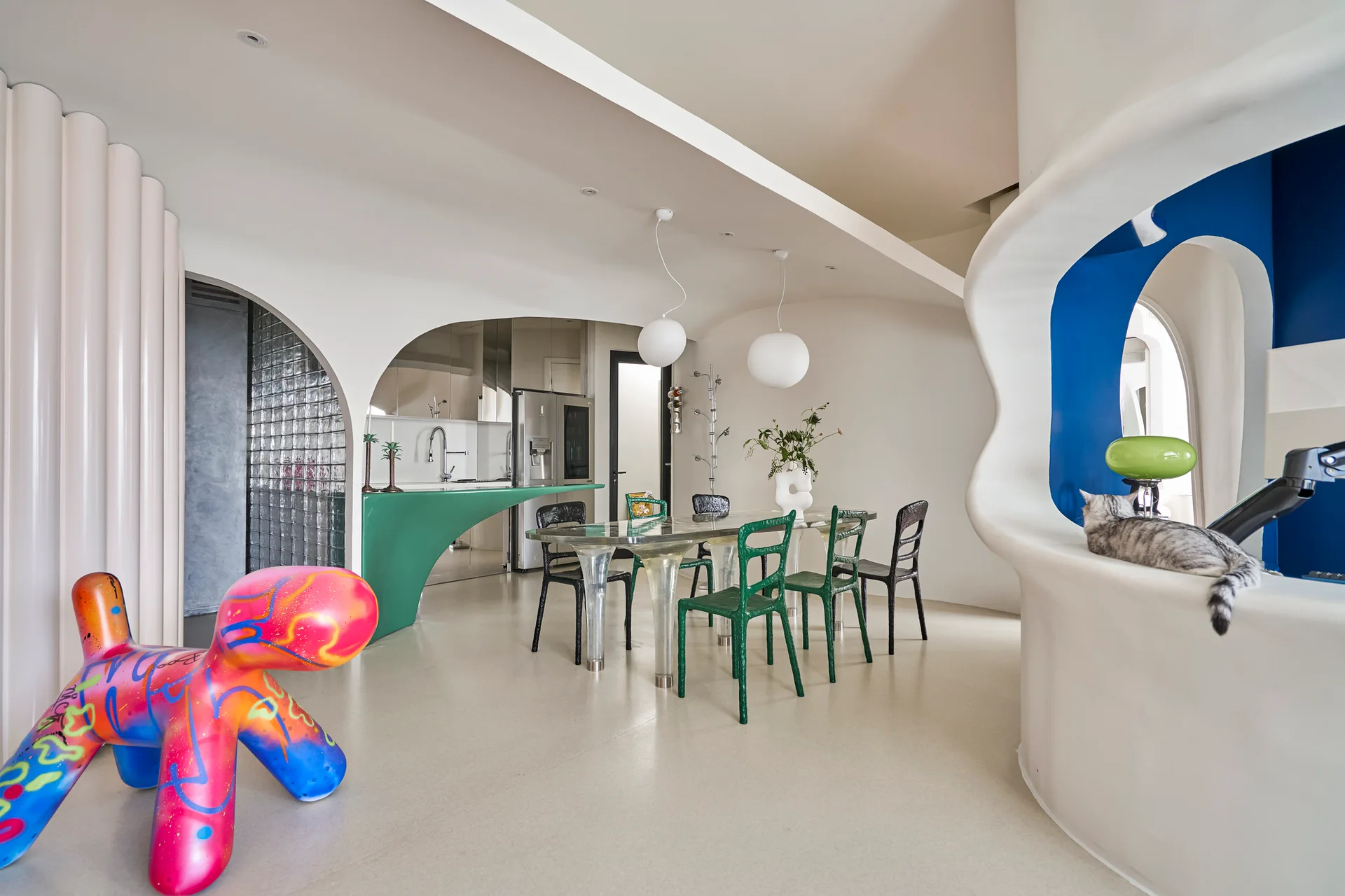 A modern dining room features organic curved archways, a transparent dining table with green chairs, and a colorful dog-like sculpture.