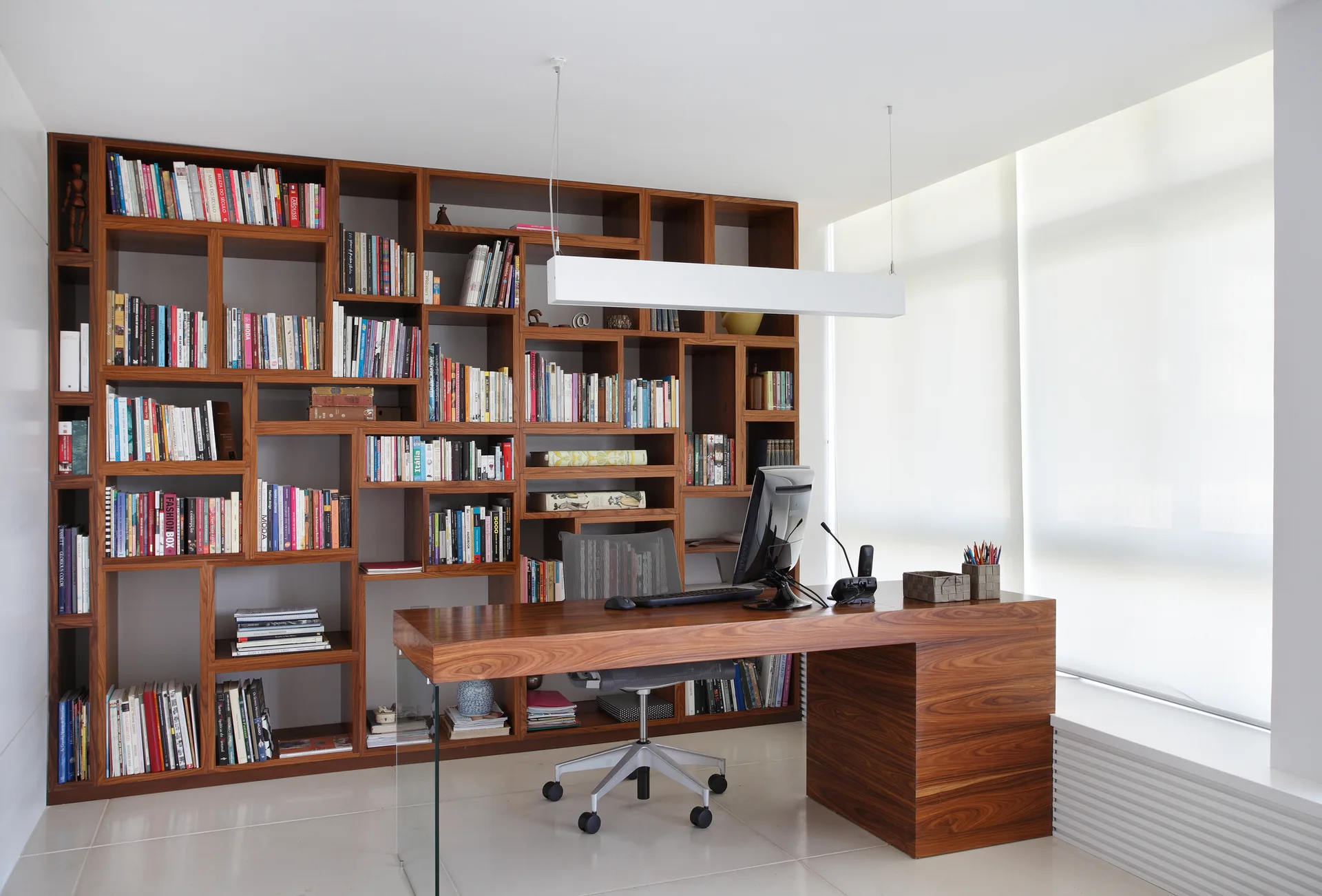 A modern home office features a wooden desk with a glass support, a mesh swivel chair, and a large floor-to-ceiling wooden bookshelf filled with books.