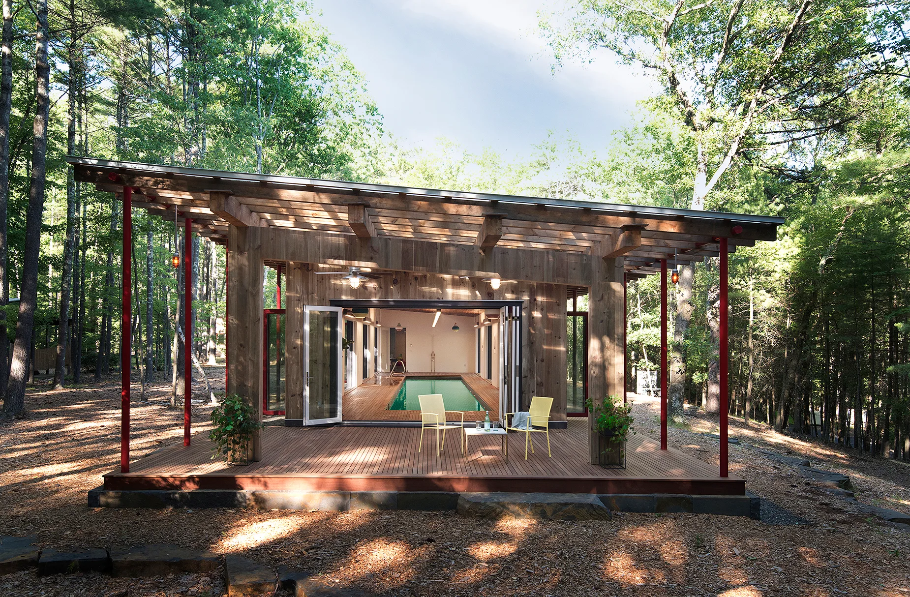 A modern wood pool house with an expansive deck, yellow chairs, and glass doors open to an indoor lap pool in a forest.