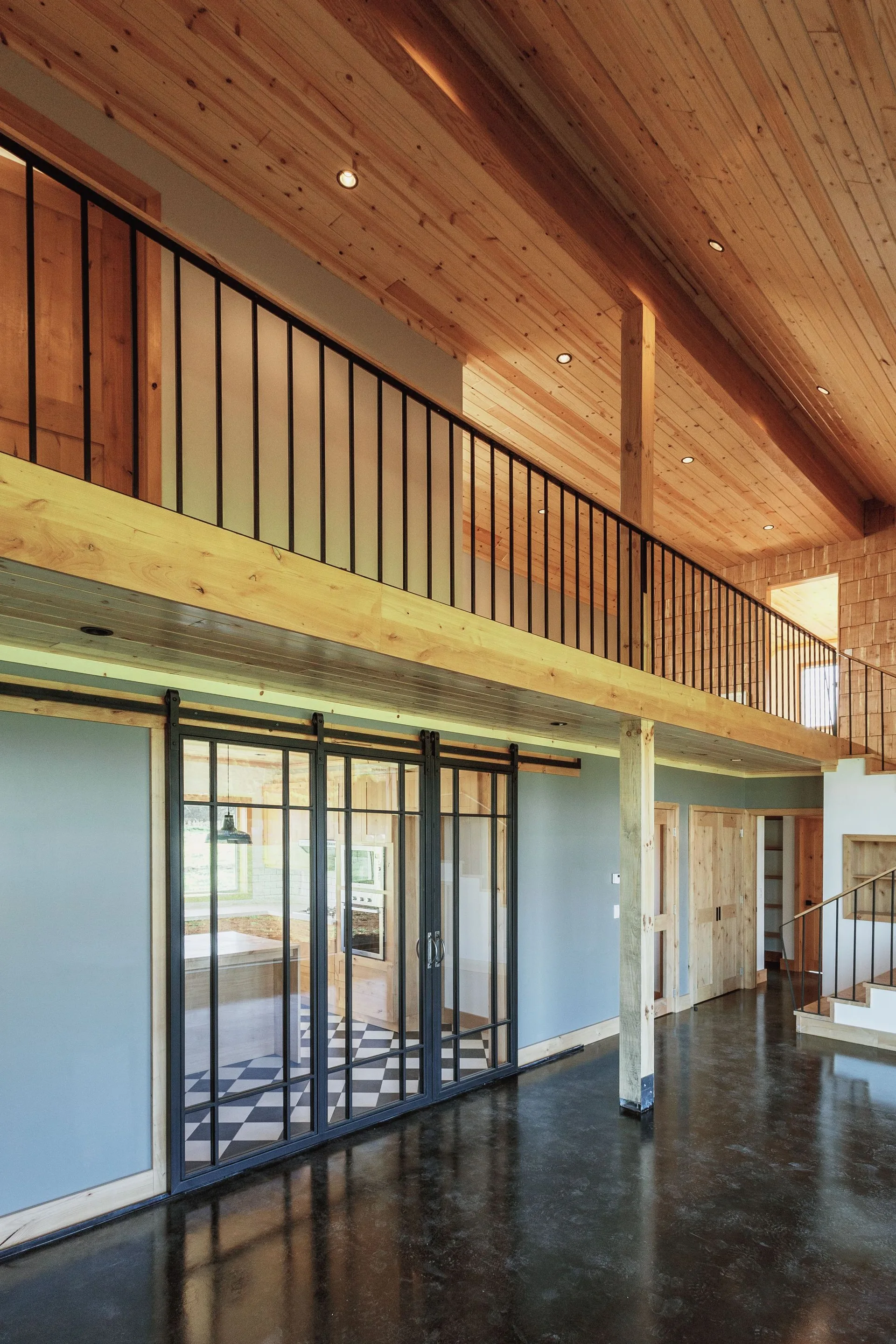 A spacious home interior features a natural wood ceiling, a mezzanine level with black metal railings, and black-framed sliding glass doors.