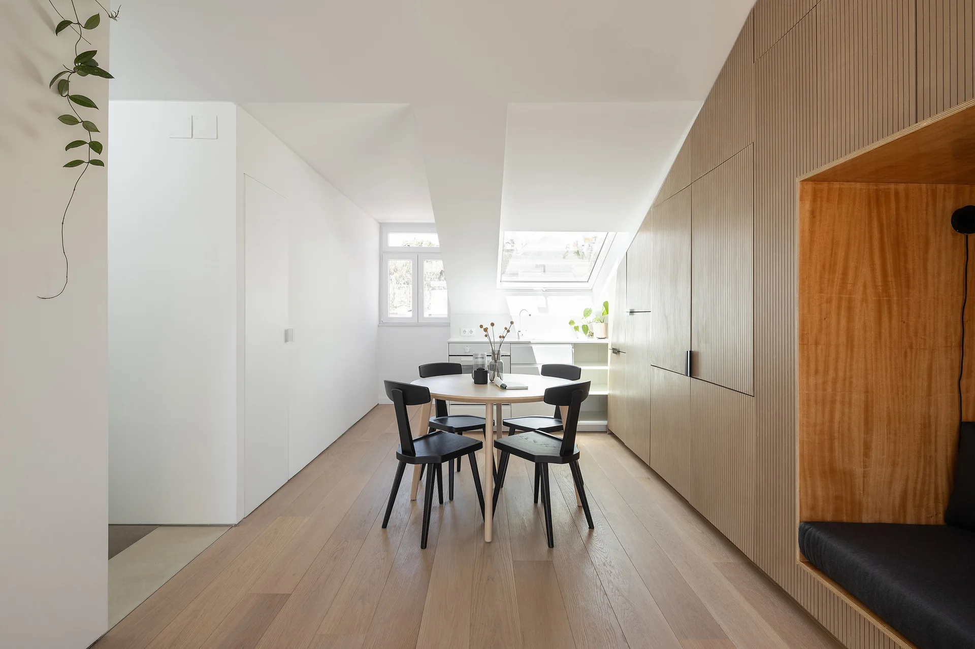 A round wood dining table and black chairs sit beneath a sloped white ceiling with a skylight next to a slatted wood storage wall.