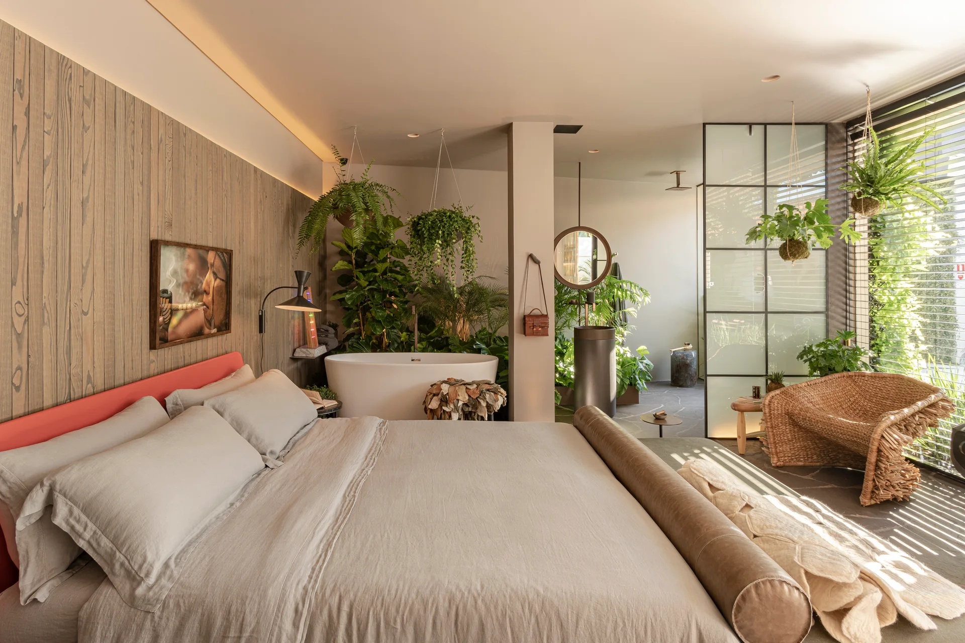A bedroom features a bed with linen sheets, vertical wood wall paneling, a freestanding bathtub surrounded by green plants, and a rattan chair.