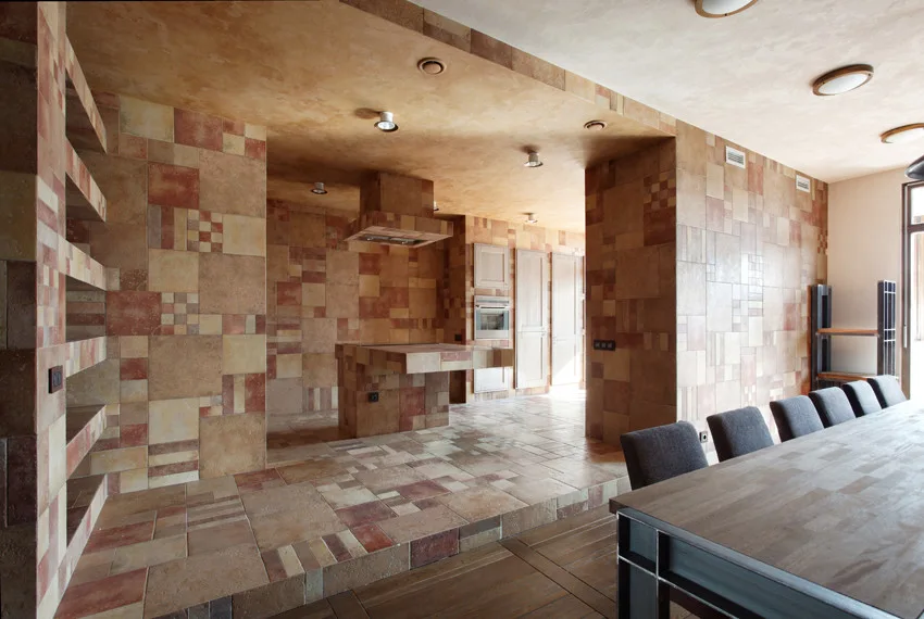 A spacious kitchen and dining area clad in earth-toned patchwork stone tiles across the walls, floor, and island, next to a long wooden dining table.