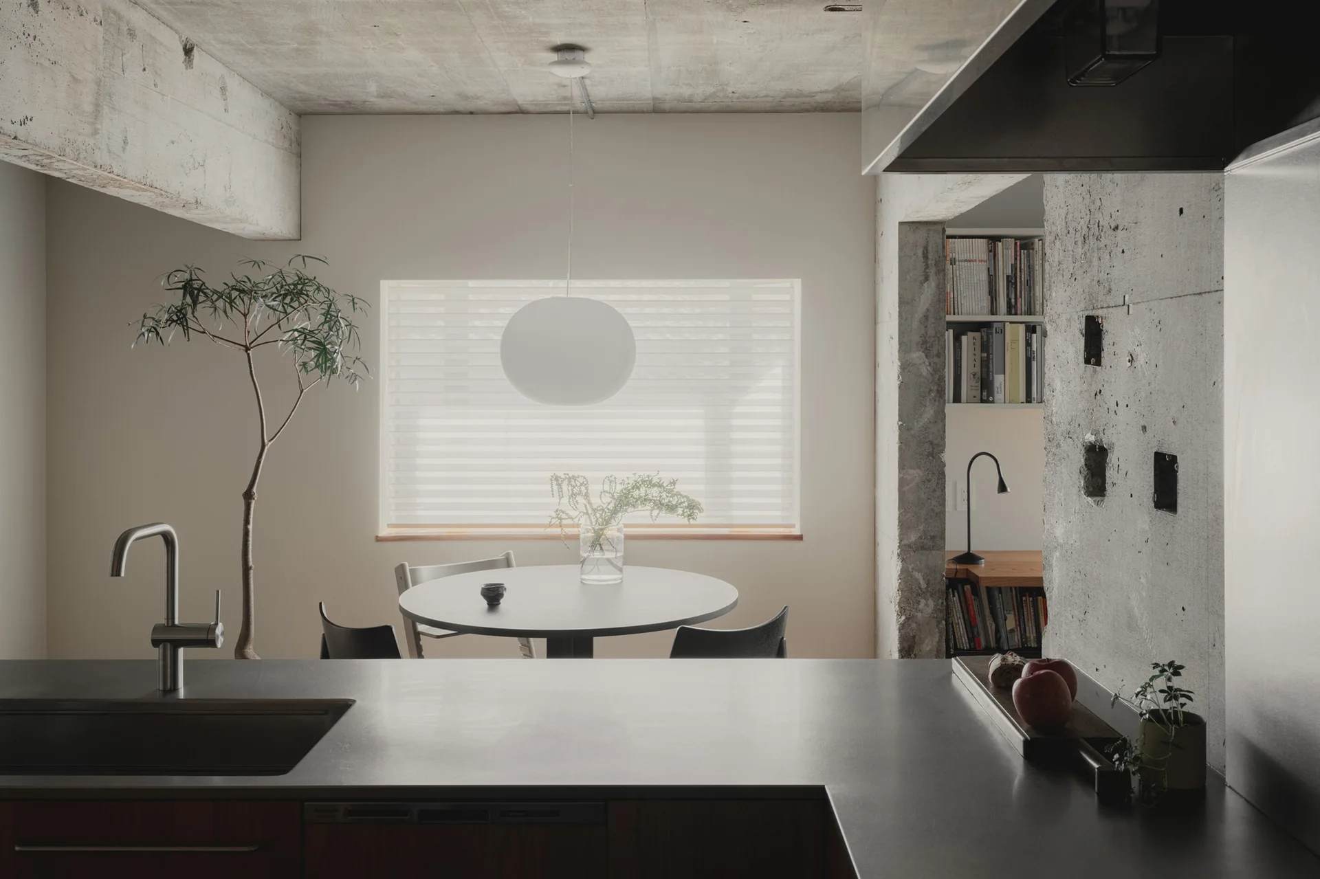 A dark kitchen counter sits in the foreground, looking toward a circular dining table under a round white pendant light with an exposed concrete ceiling.