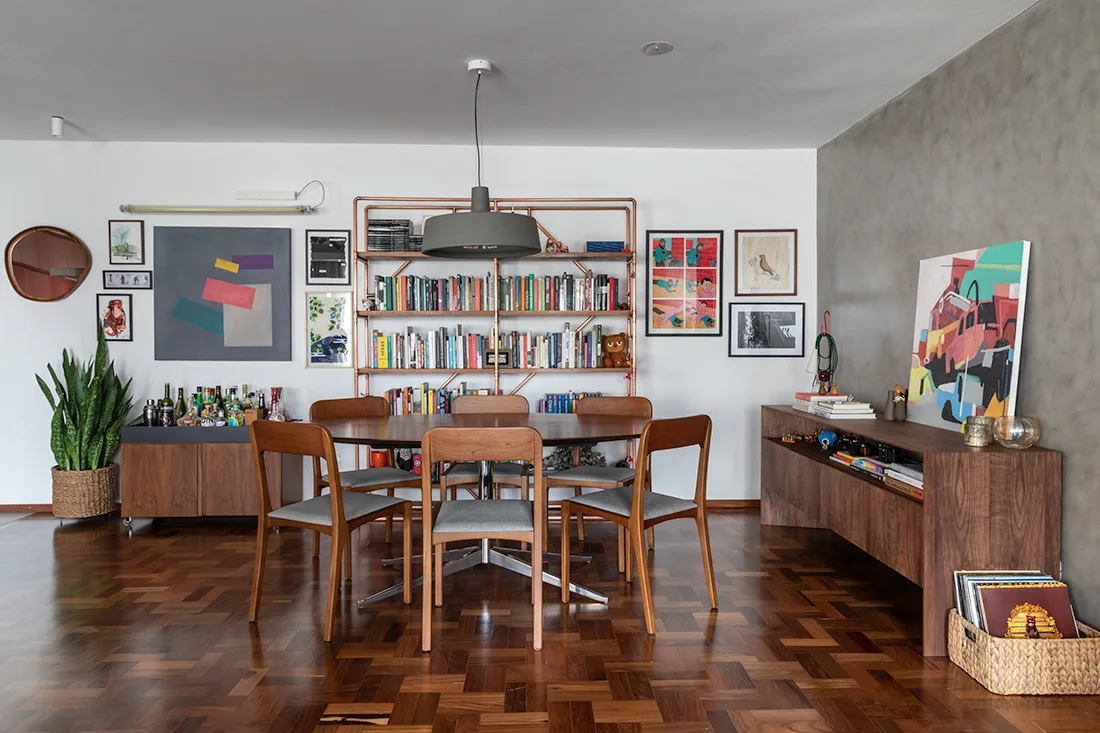 A mid-century dining room features an oval wooden table on parquet flooring, accented by a copper pipe bookshelf and a grey textured wall.