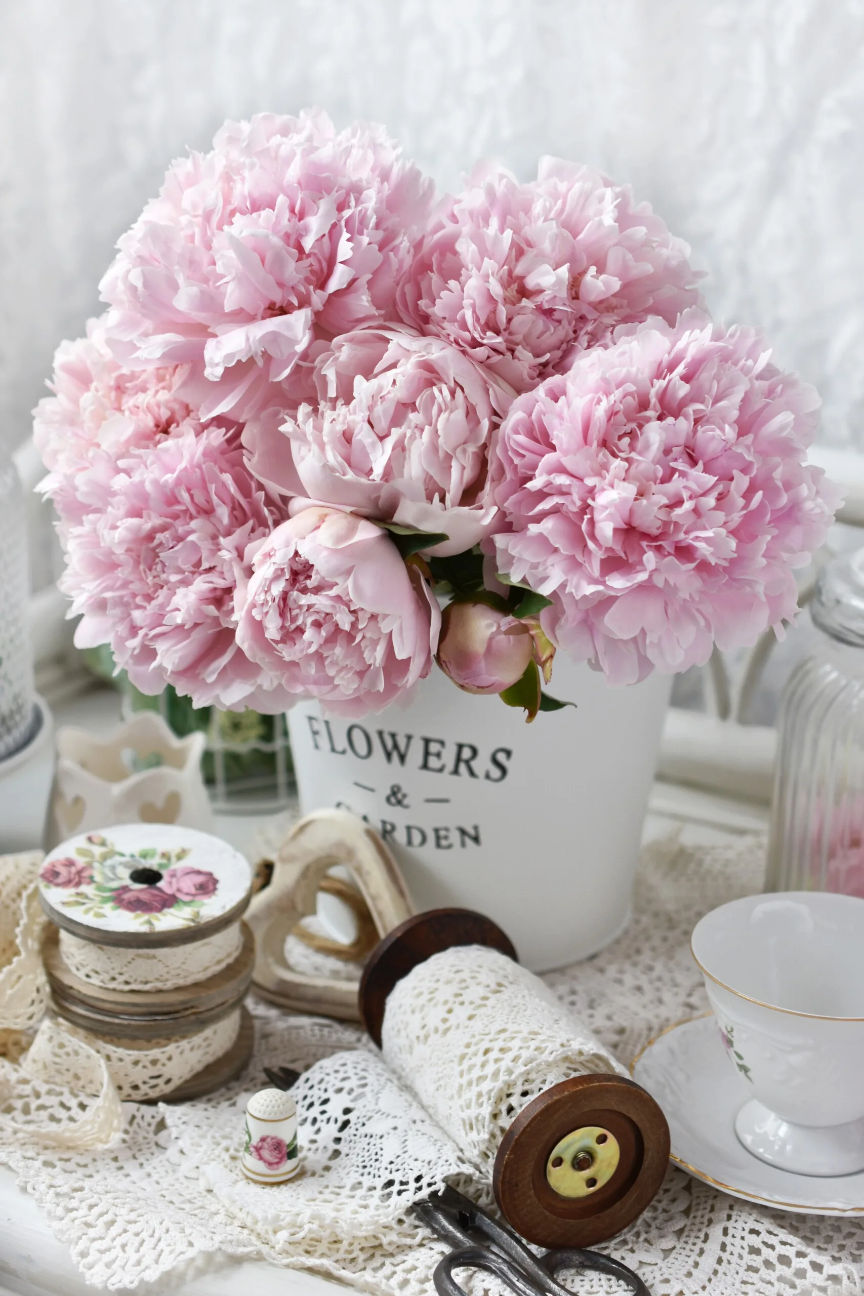 A close-up of a tabletop display featuring pink peonies in a white planter alongside vintage wooden lace spools, a heart ornament, and a porcelain teacup.