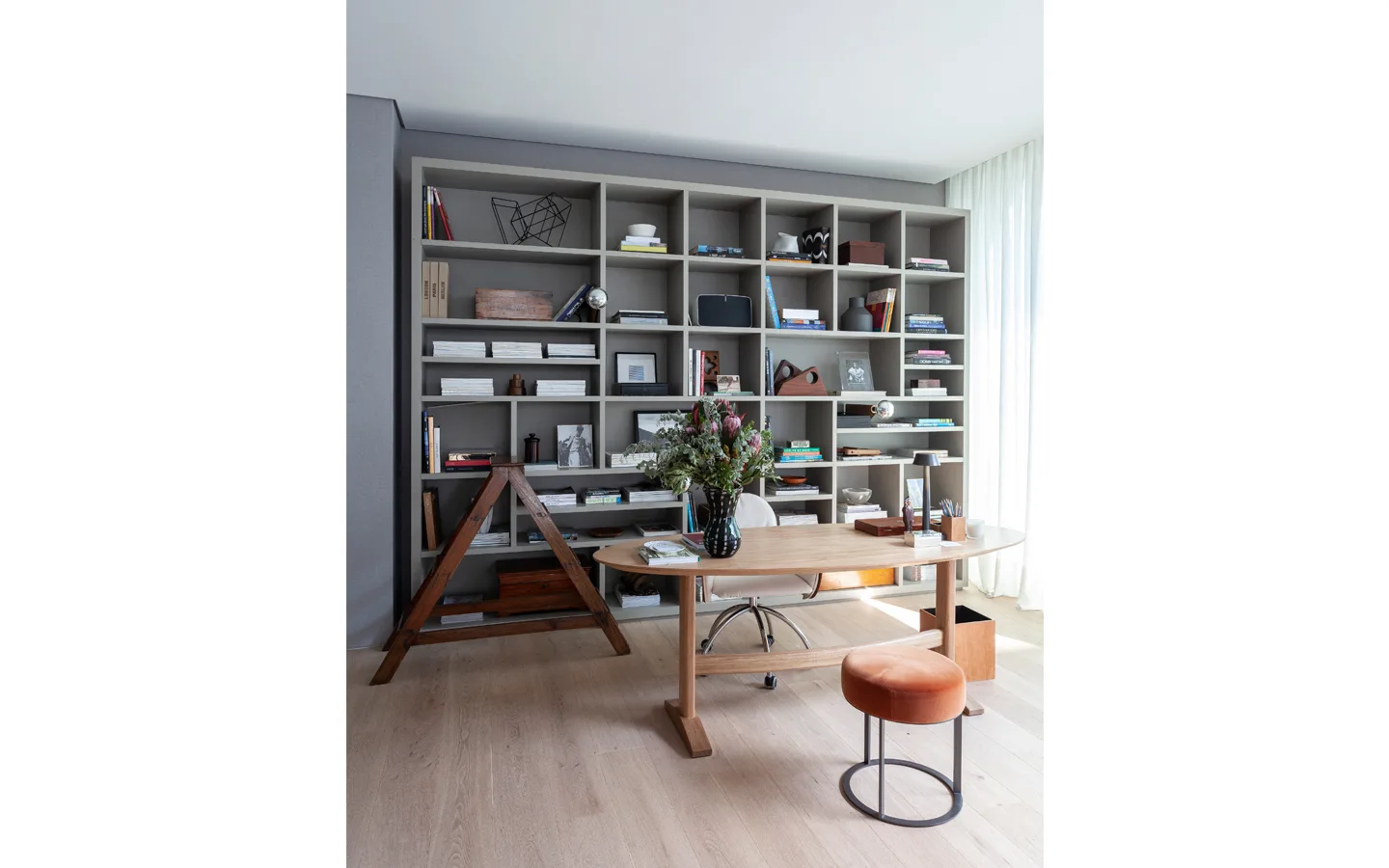 A home office features a large grey gridded bookshelf against the wall, a light wood oval desk with an orange stool, and a swivel chair.
