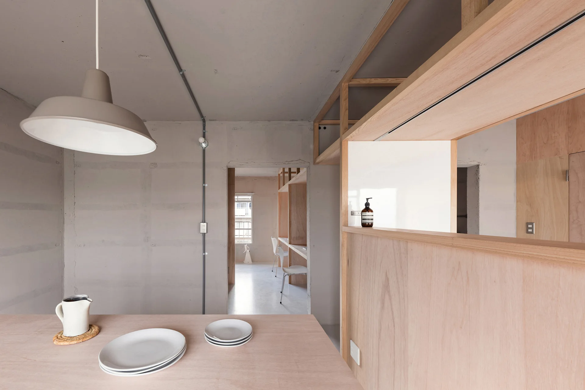 A minimalist dining space features a light wood table, a beige pendant light, exposed concrete walls, and a plywood partition wall.