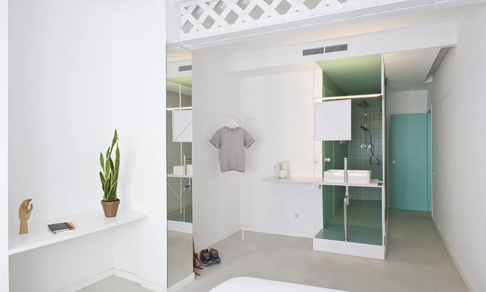An open-concept minimalist bedroom and bathroom featuring white walls, a seafoam green tiled shower stall, a vertical mirror, and a low floating shelf.