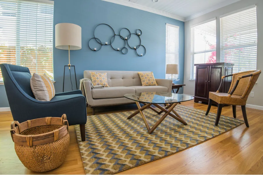 A living room with a blue accent wall, light grey sofa, navy blue armchair, glass coffee table, and a geometric patterned area rug.