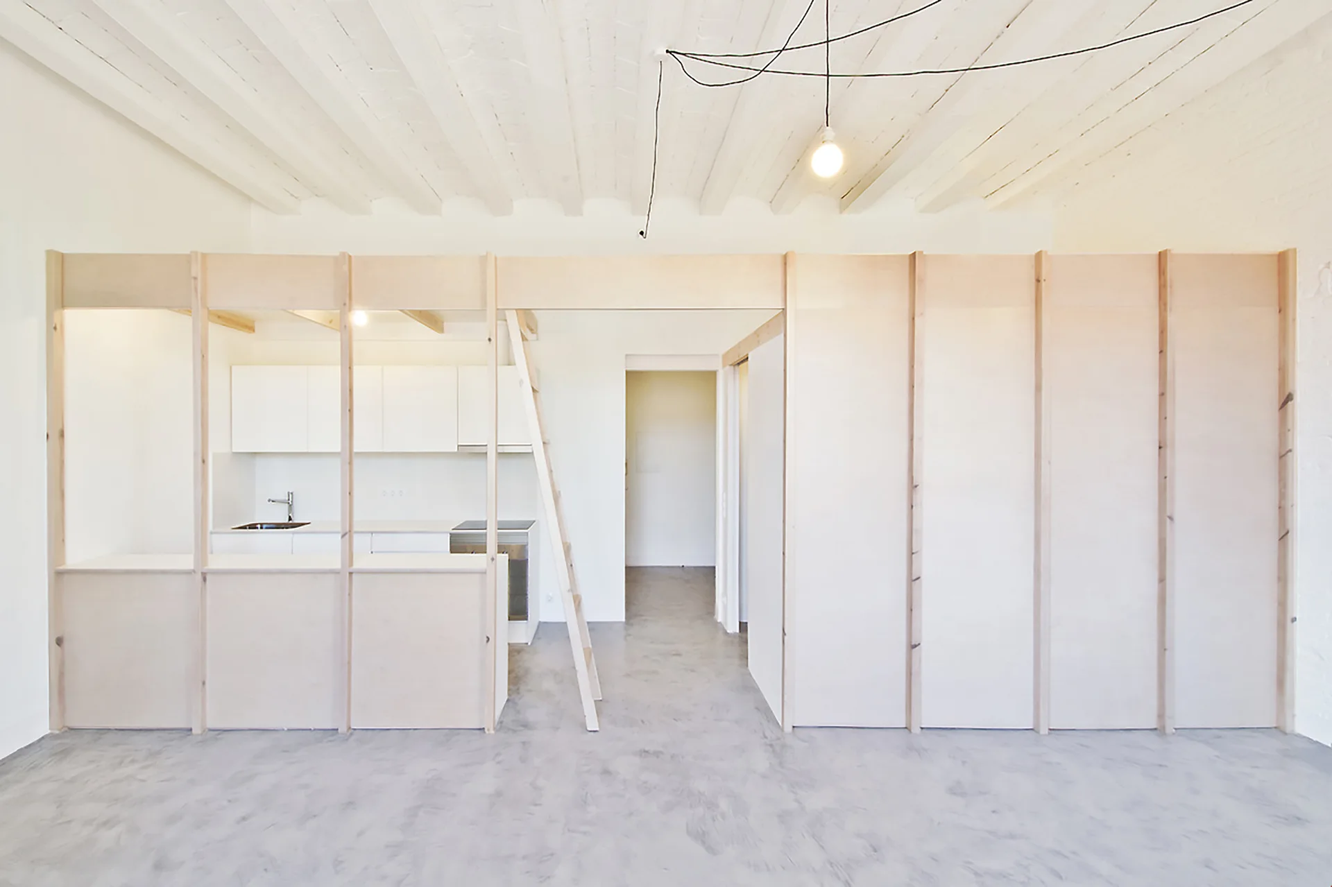 A minimalist studio features a light wood plywood partition wall separating an open kitchen from a polished grey floor under painted ceiling beams.