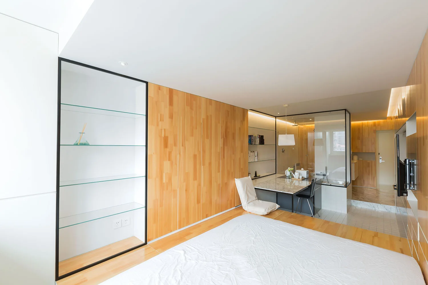 A minimalist bedroom features light wood wall paneling, a bed in the foreground, and a glass-enclosed bathroom next to a marble-topped desk.