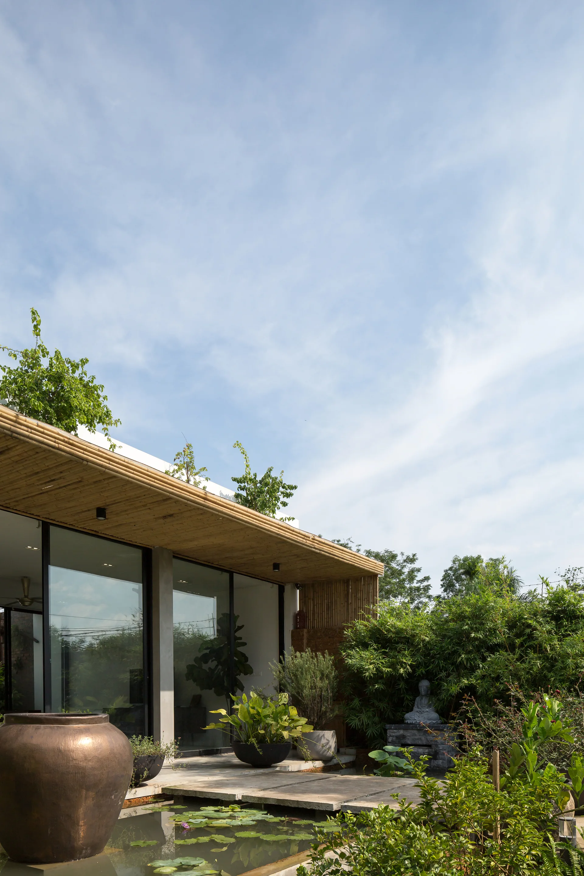A modern tropical house facade with floor-to-ceiling glass doors, a textured bamboo overhang, and a concrete stepping stone path crossing a lily pond.