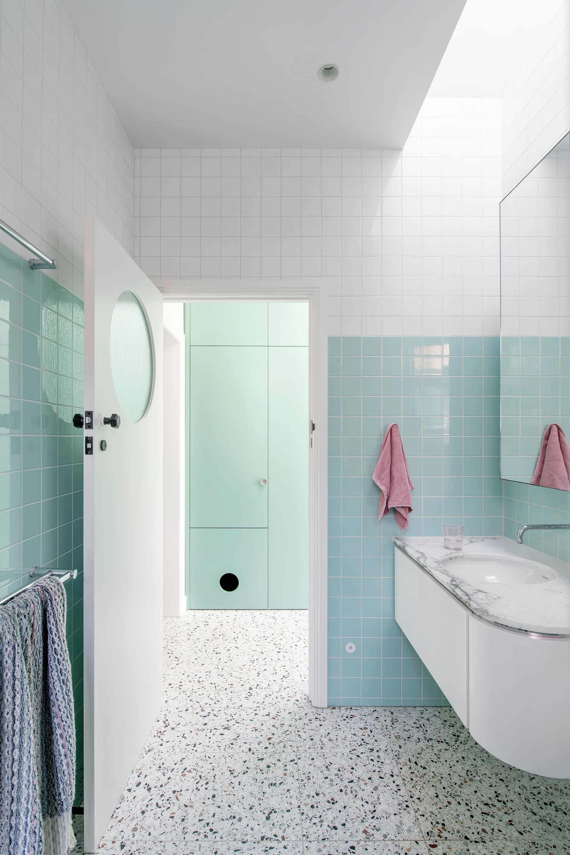 A contemporary bathroom features pale blue square tiles, a white floating vanity with a marble top, and speckled terrazzo flooring under a skylight.