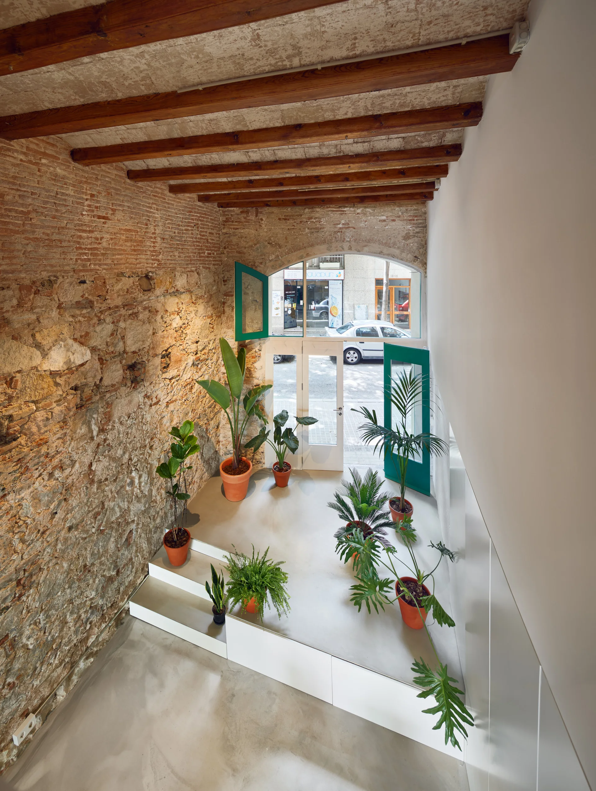 An entryway seen from above features an exposed brick wall, wooden ceiling beams, and green potted plants on a tiered white platform.