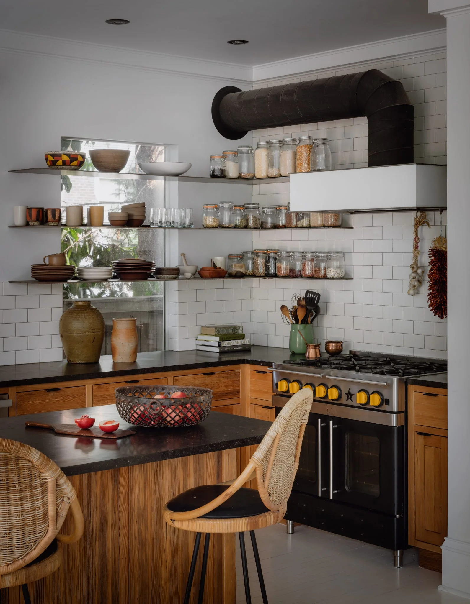 A kitchen with warm wood cabinets, open steel shelving holding glass jars, white subway tile backsplash, and two rattan stools at a dark island.