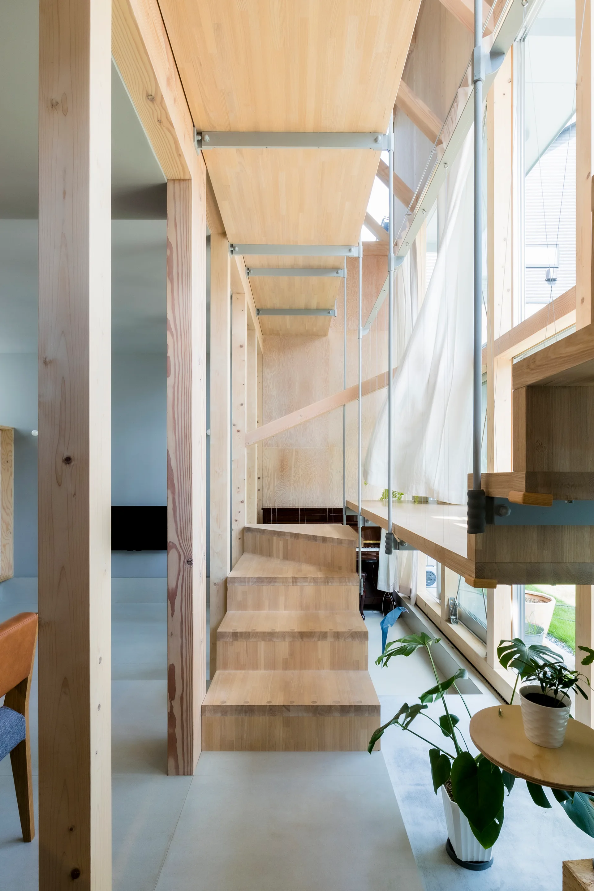 A central light wood staircase rises through an exposed timber frame interior beside a large window with sheer white curtains.