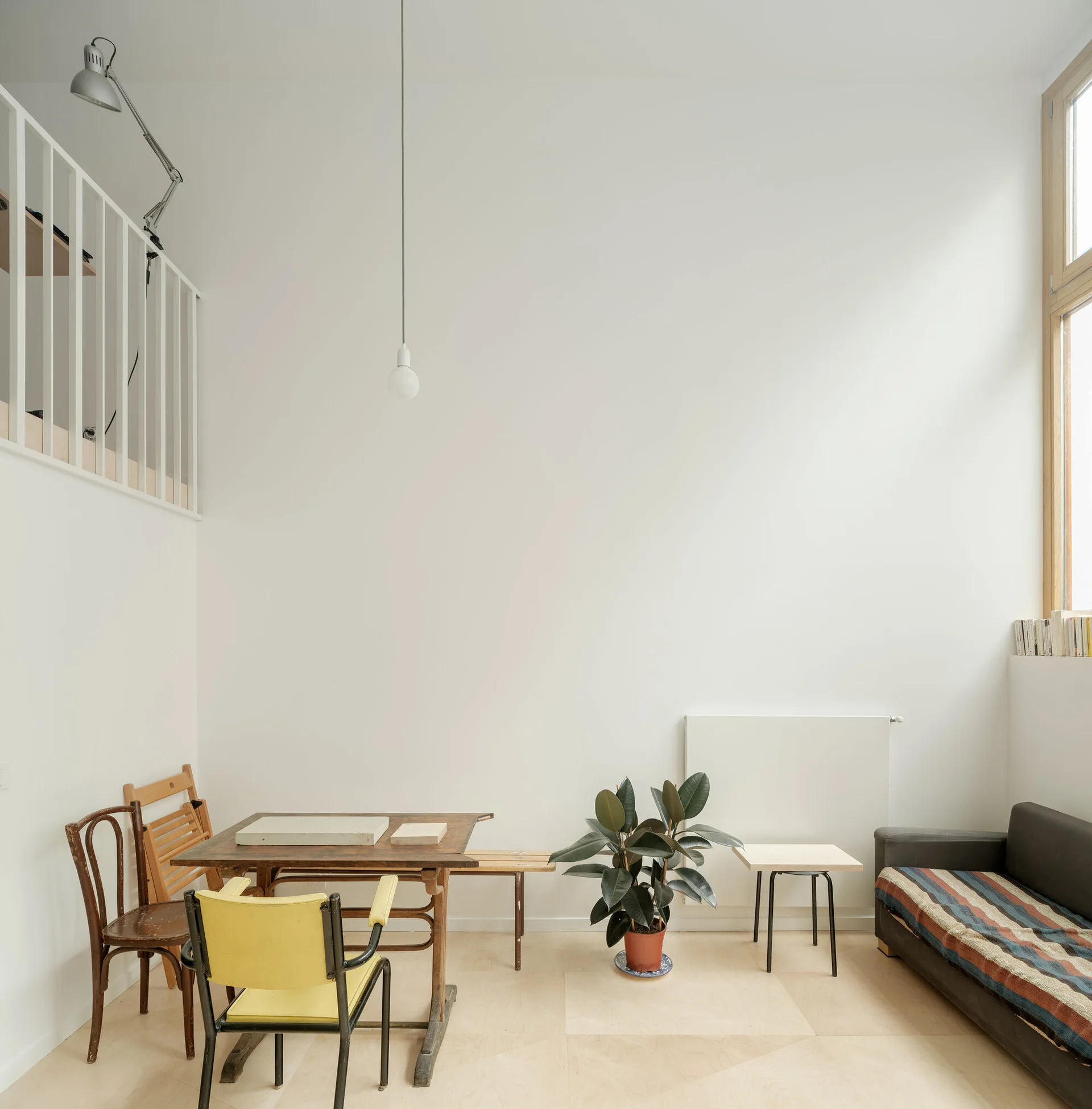A minimalist living area with a high ceiling, white walls, a wooden dining table with a yellow chair, a potted plant, and a dark sofa.