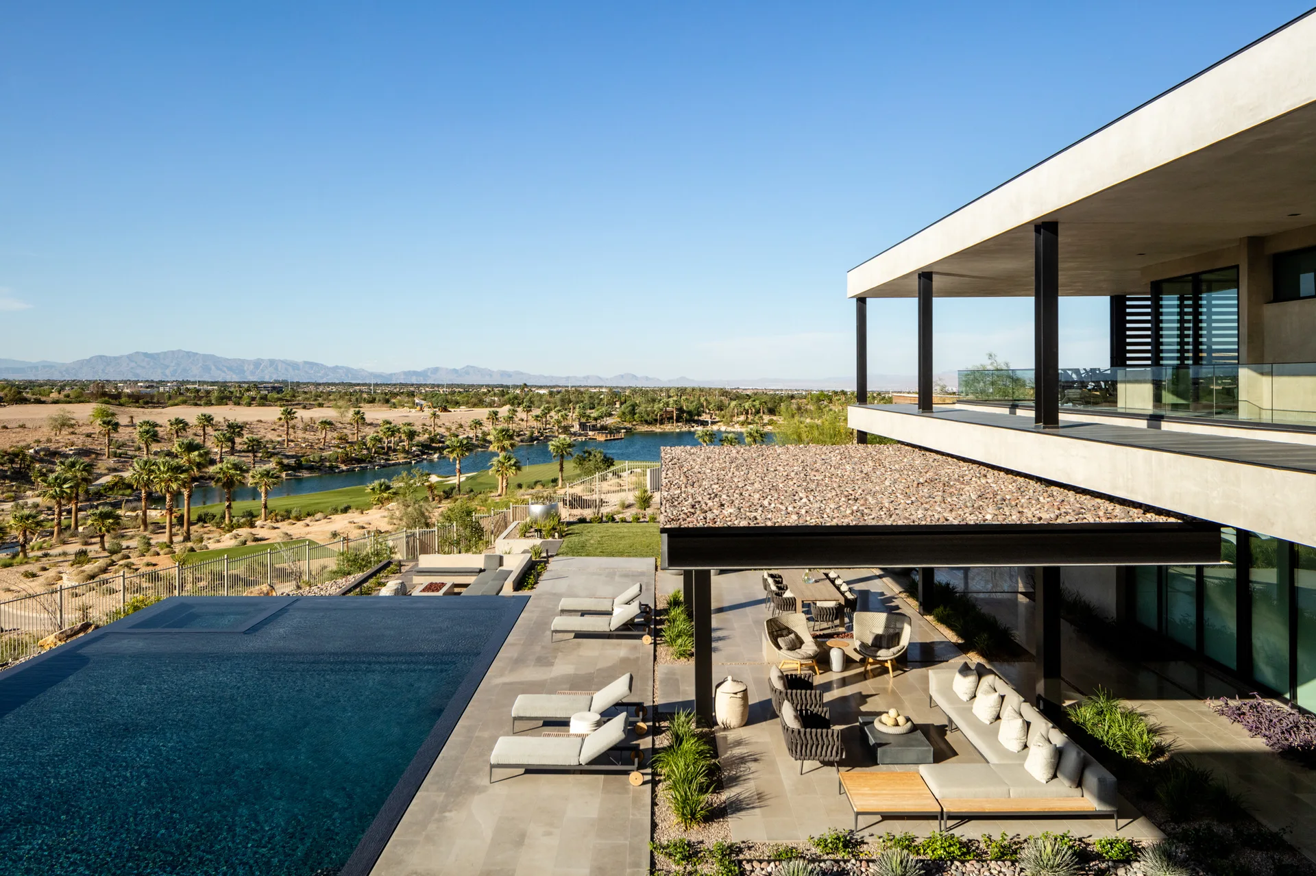 A modern multi-level patio features a rectangular infinity pool, grey stone pavers, and a covered lounge area with a sectional sofa overlooking palm trees.