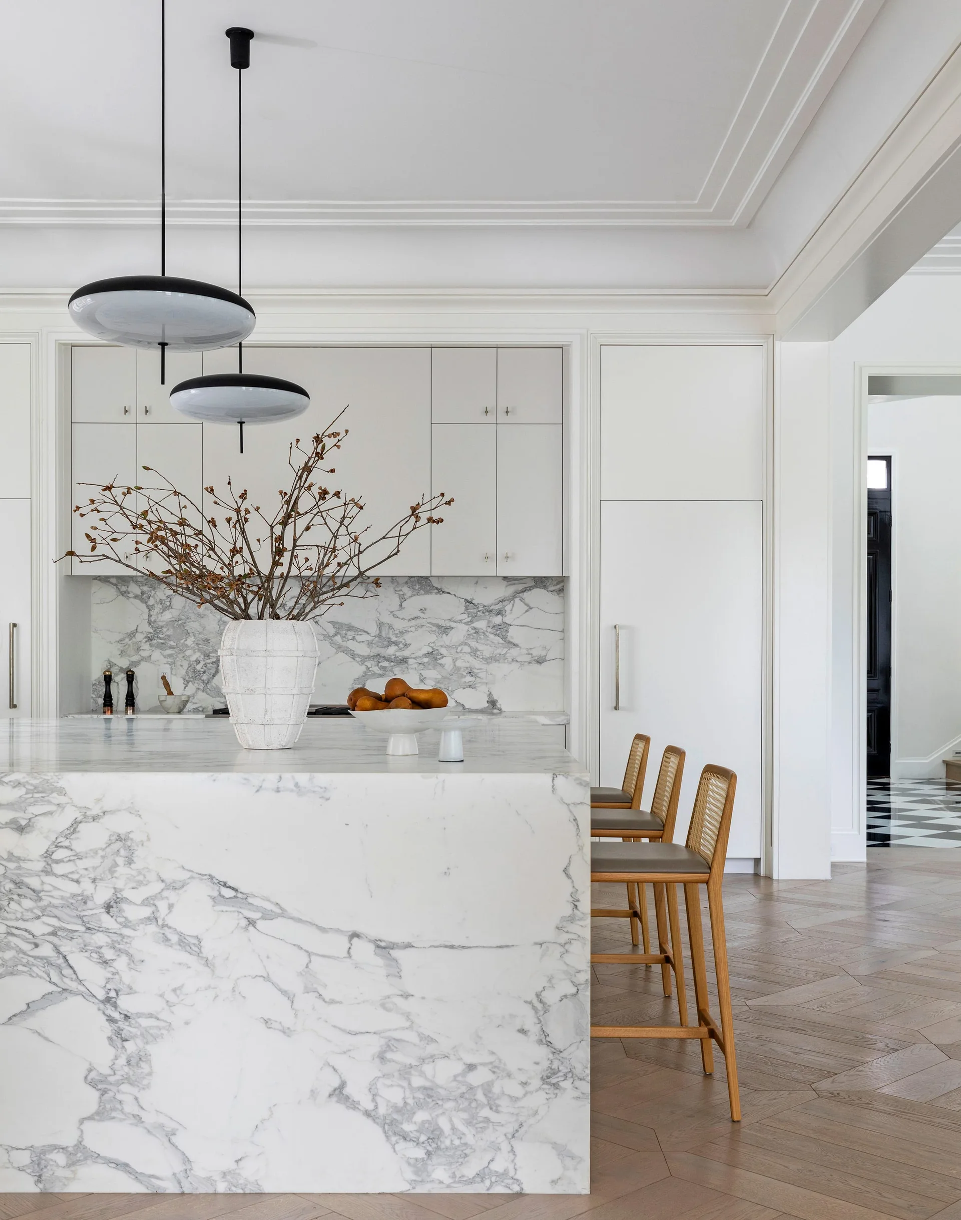 A marble kitchen island with a matching backsplash features rattan bar stools, integrated white cabinetry, and a wood herringbone floor.
