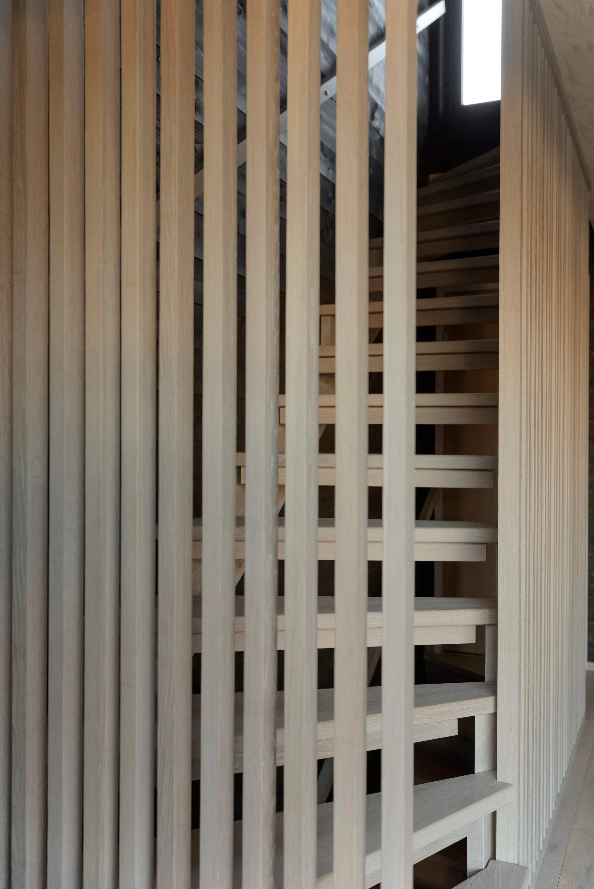 A minimalist light wood staircase featuring open treads is viewed through a screen of floor-to-ceiling vertical wooden slats.