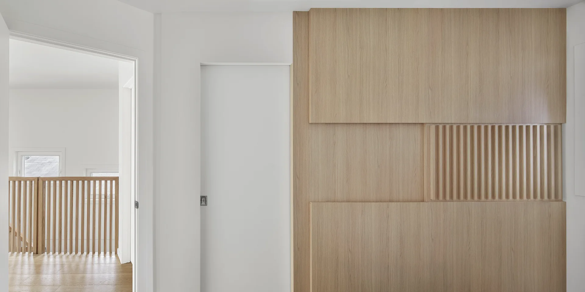A minimalist white hallway featuring a flush sliding door next to a large light wood accent wall with integrated fluted vertical paneling.